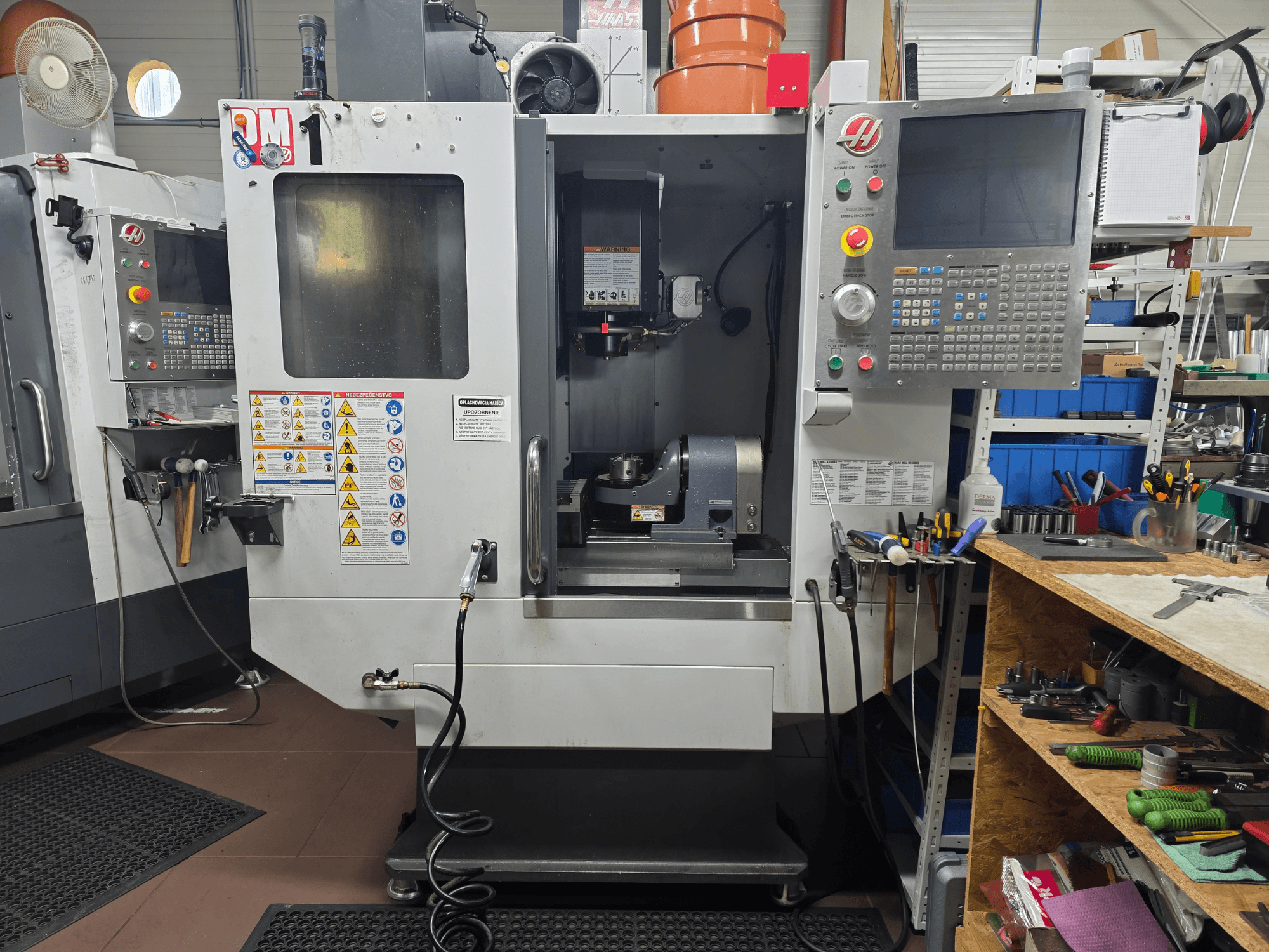 Haas CNC machine model VF-2, front view, showing controls, spindle area, safety labels, and tools on a nearby workbench.