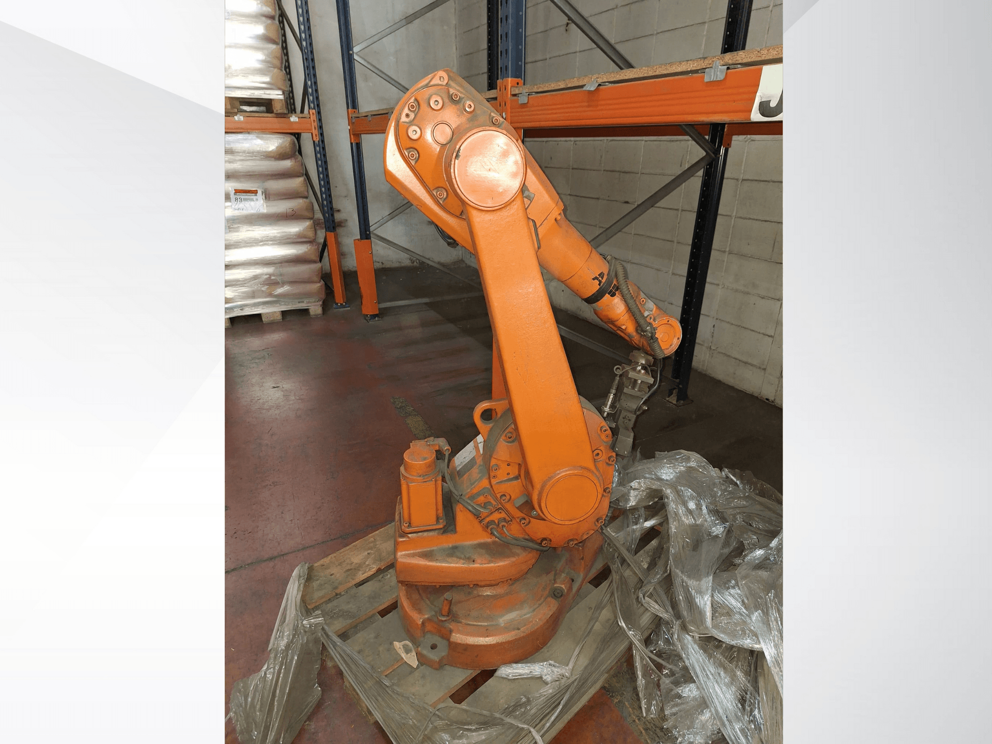 Industrial robotic arm in orange, front view on wooden pallet, with attached cable and protective plastic wrap around base.