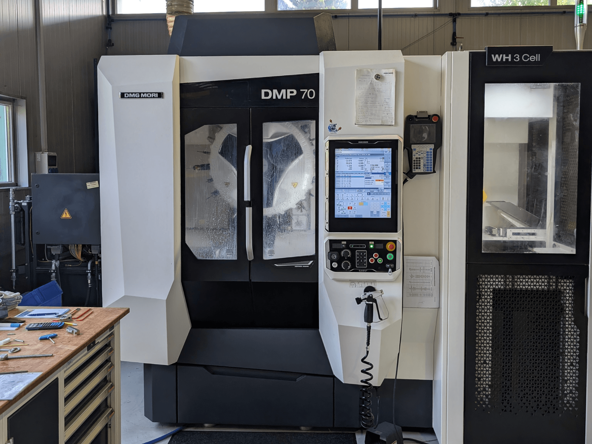 Vertical machining center DMG MORI DMP 70, front view with control panel and operational display, surrounded by workshop tools.