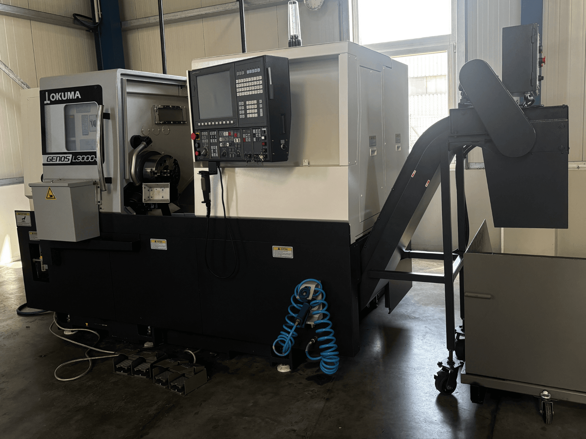 Okuma GENOS L3000-e horizontal turning machine, front view, features control panel, conveyor, and visible spindle area.