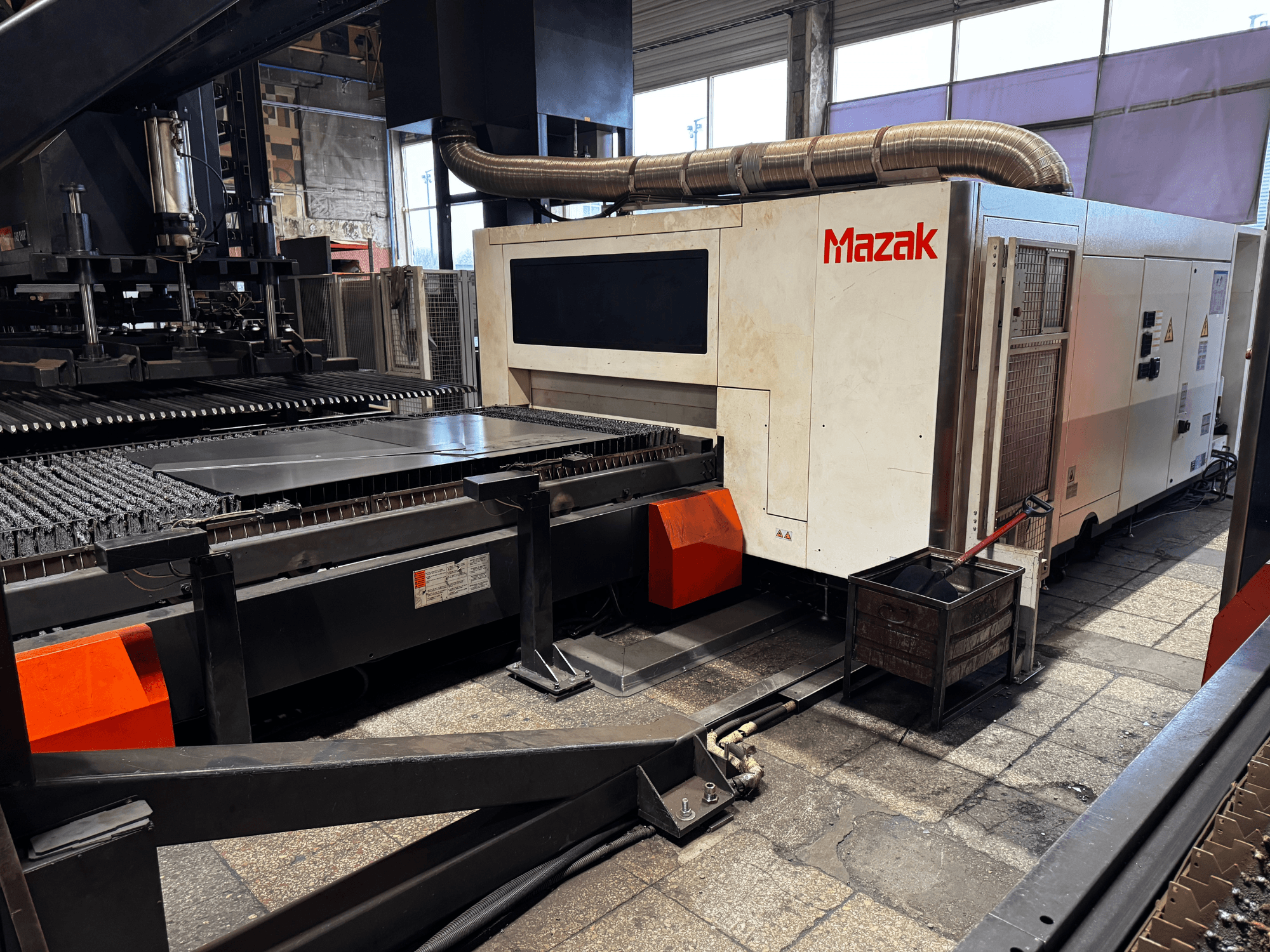 Mazak laser cutting machine in a workshop, side view showing control panel, conveyor, and a collection bin.