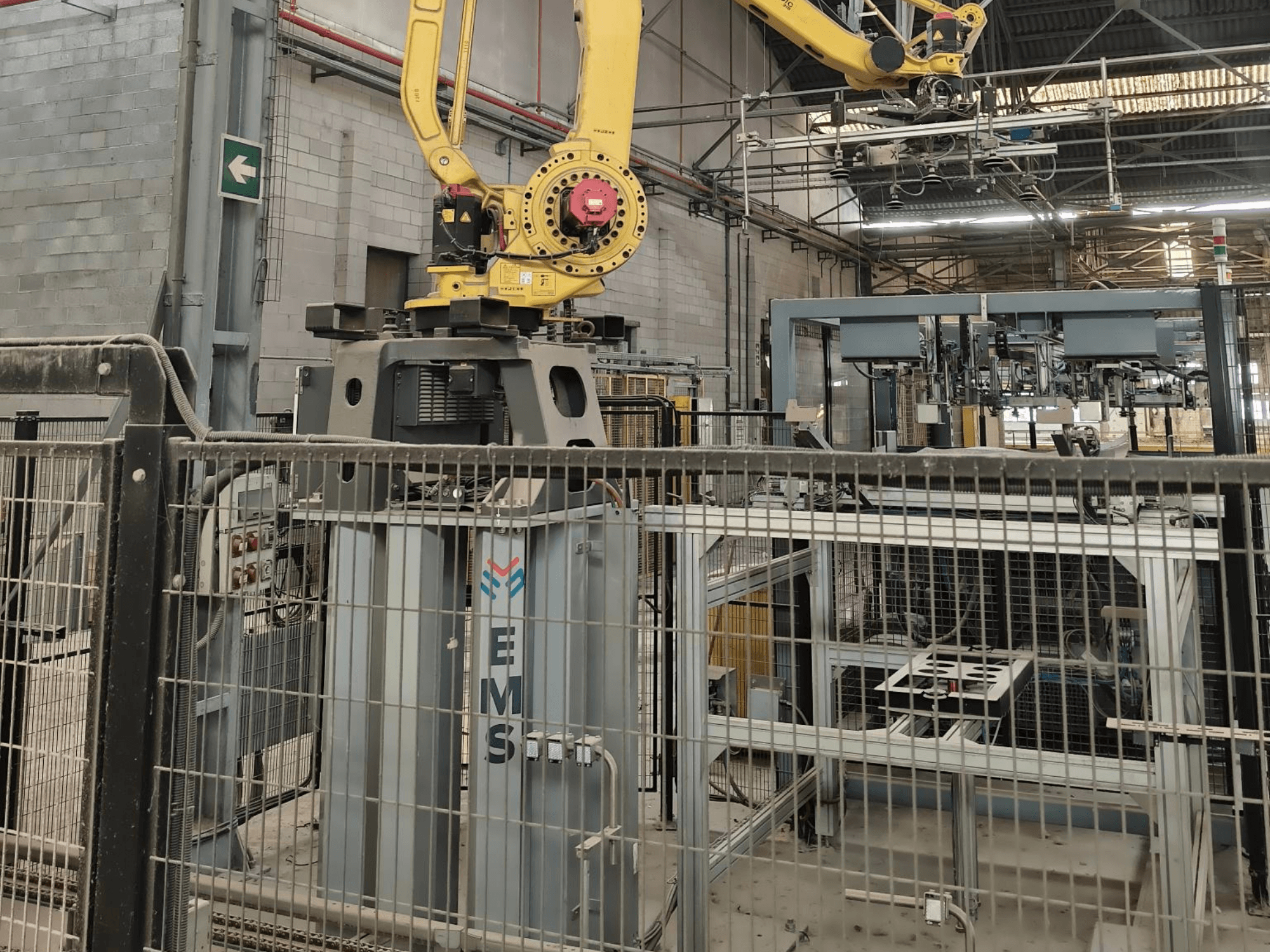 Front view of FANUC M-410iC/315 robot arm positioned over an EMS tool, with industrial equipment and fencing in the background.