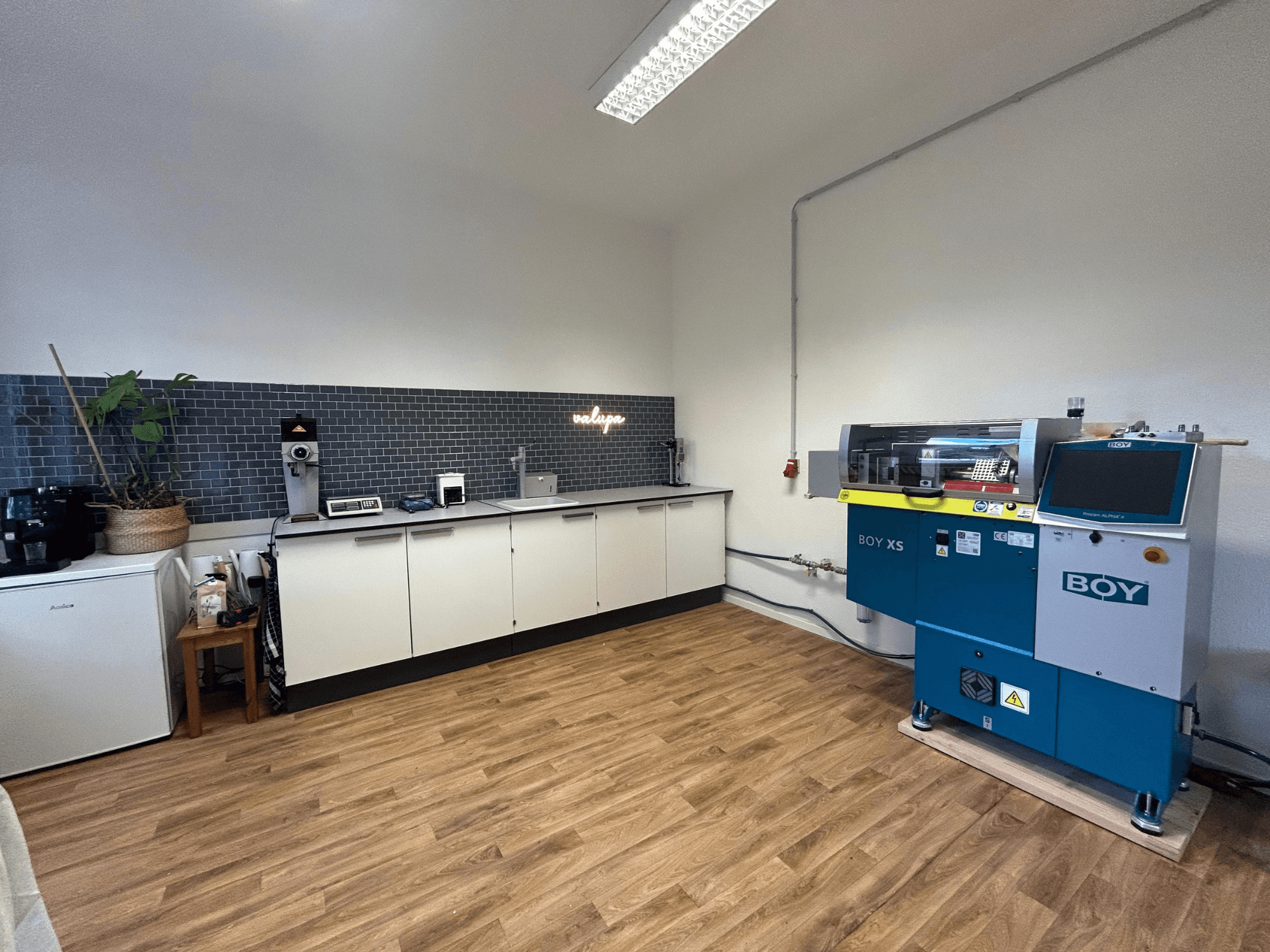 Hydraulic injection moulding machine BOY XS 161, front view, in a modern kitchen setting with appliances and wooden flooring.