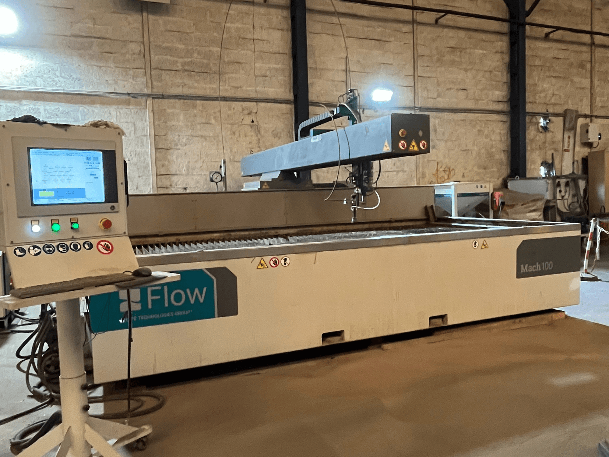 Flow Mach 100 waterjet cutter, side view, with a digital control panel and cutting head visible on a worktable.