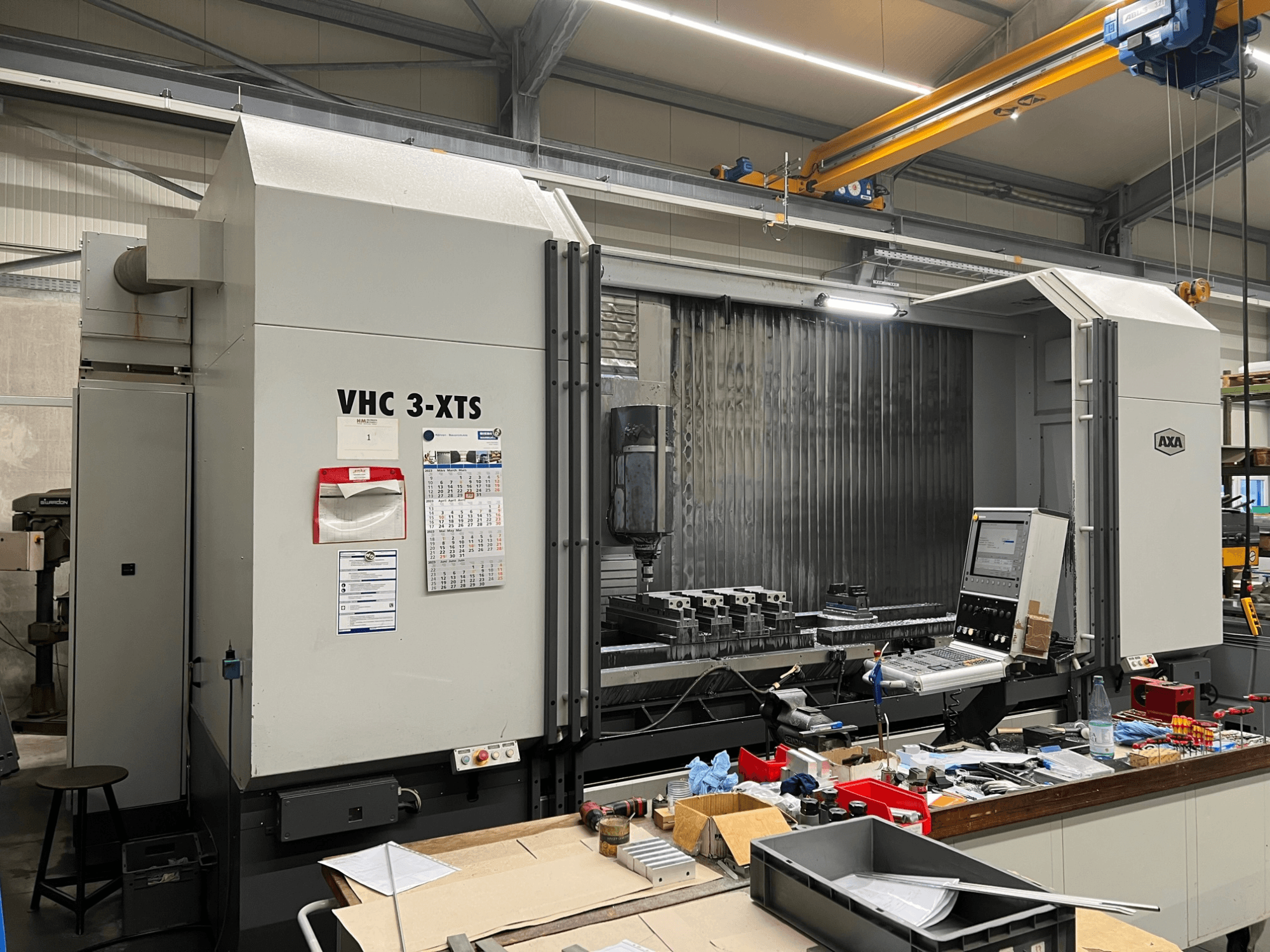 AXA VHC 3-XTS CNC machine in side view, with control panel and work area cluttered with tools and components.