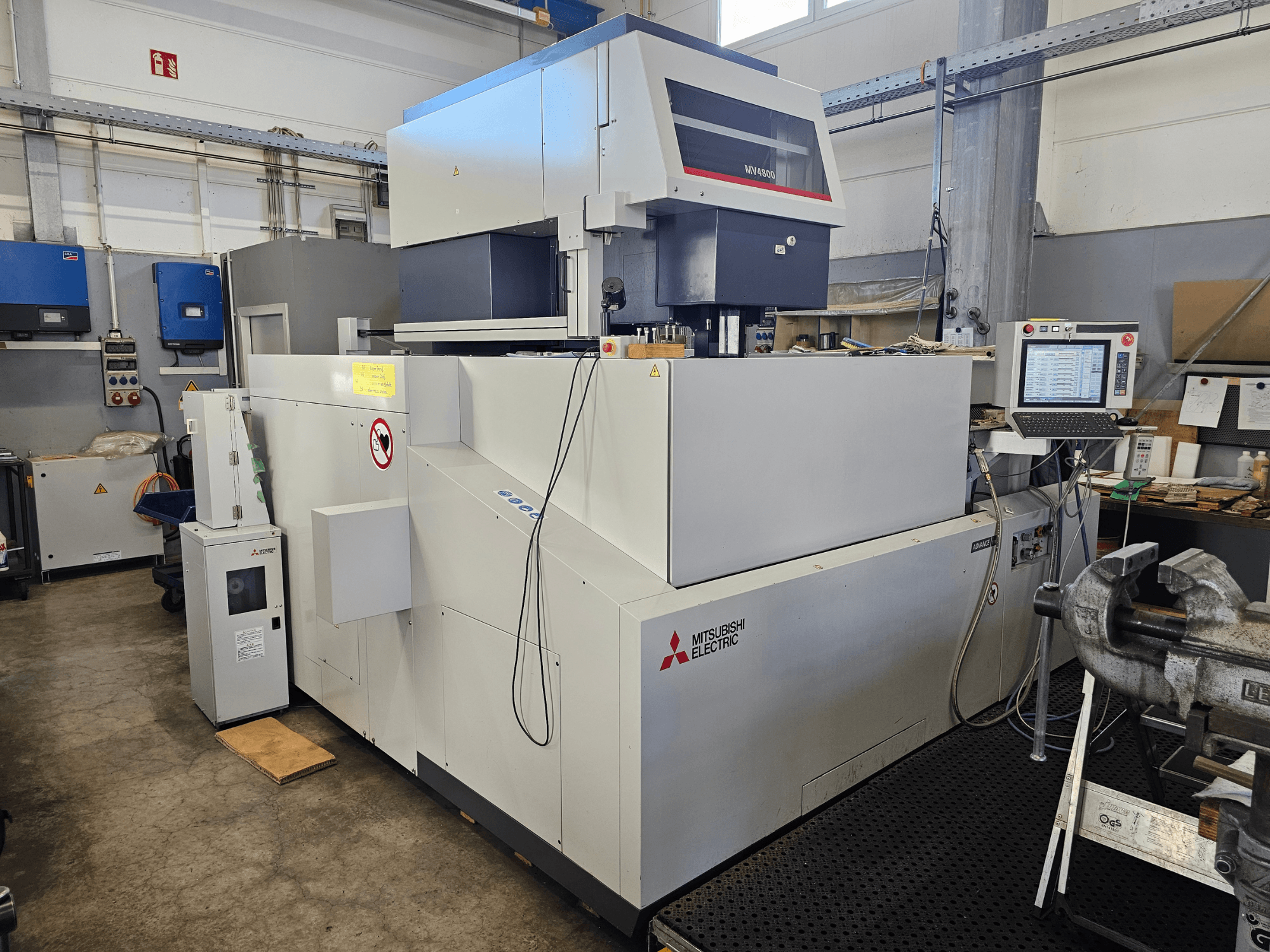 Mitsubishi MV4800 Wire EDM Machine in a workshop, side view showing control panel, safety labels, and surrounding equipment.