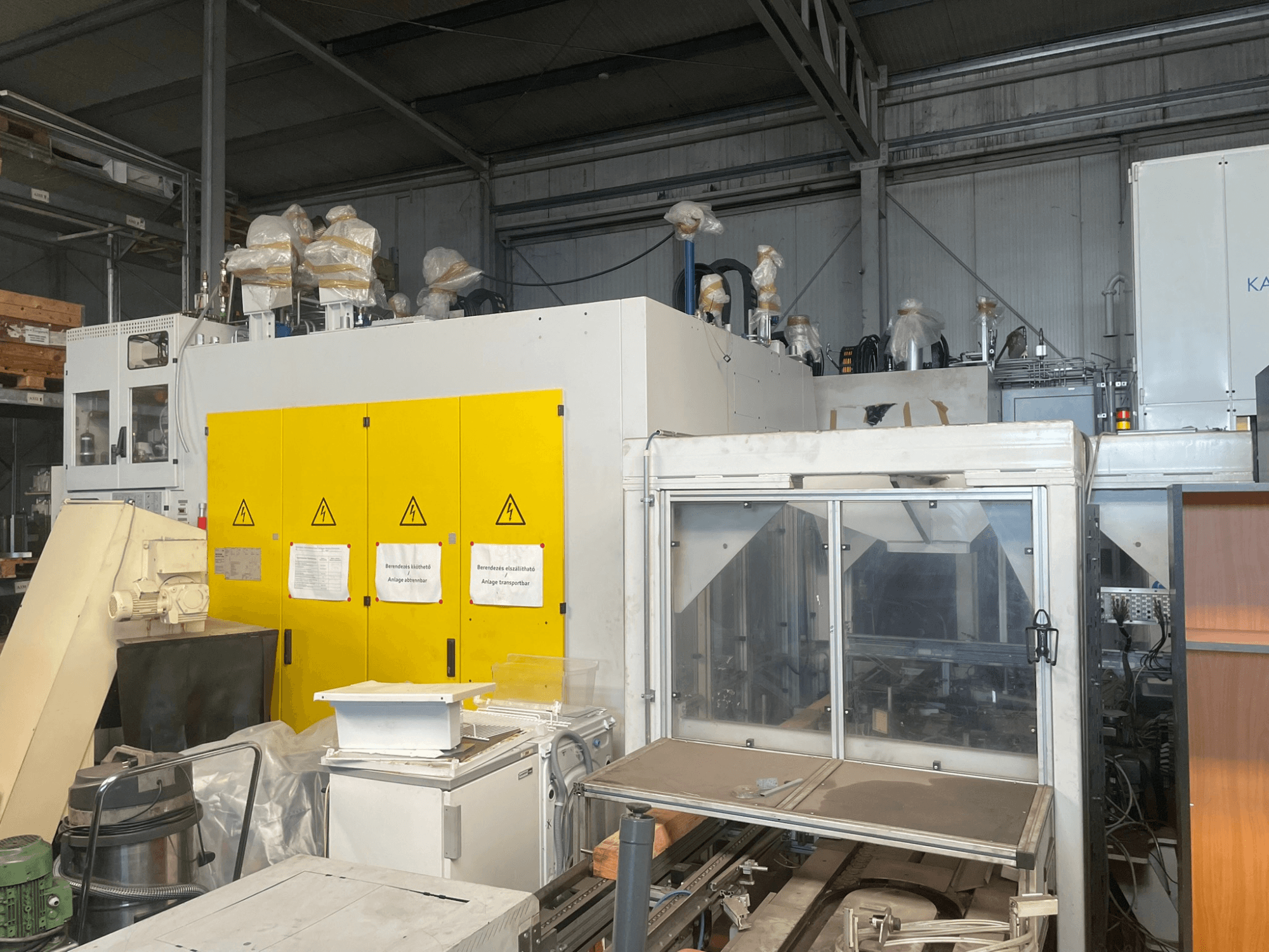 Industrial machinery with yellow control panel, side view, featuring warning labels and plastic coverings, in a warehouse setting.