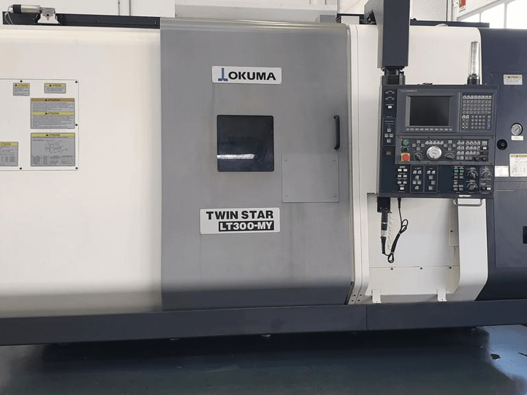 Okuma Twin Star LT300-MY CNC lathe in side view, featuring control panel, operational buttons, and safety labels.