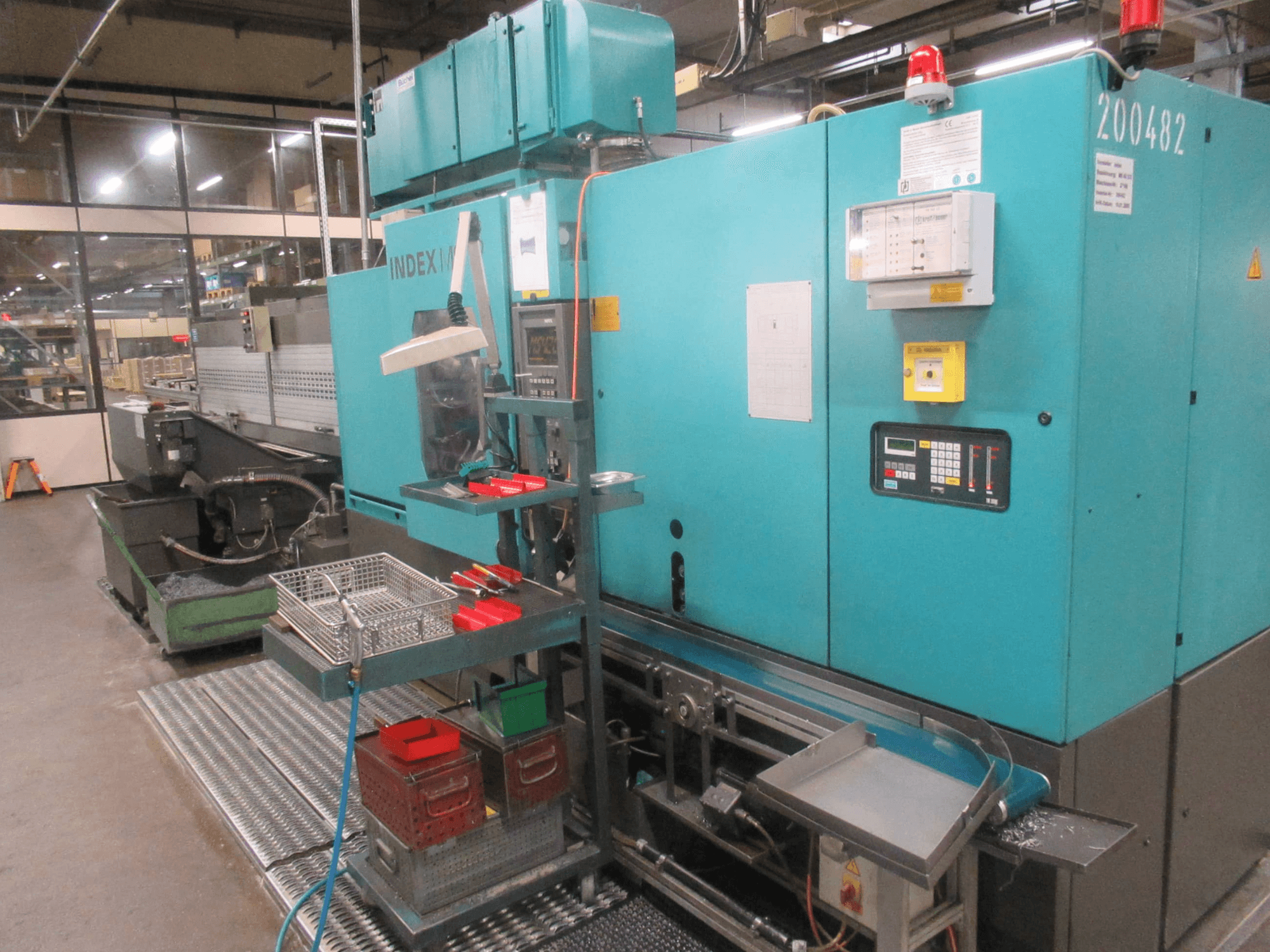 Turquoise INDEX machine in factory, side view; features control panel, tool storage, and conveyor. Visible bins, hoses, and alarms.