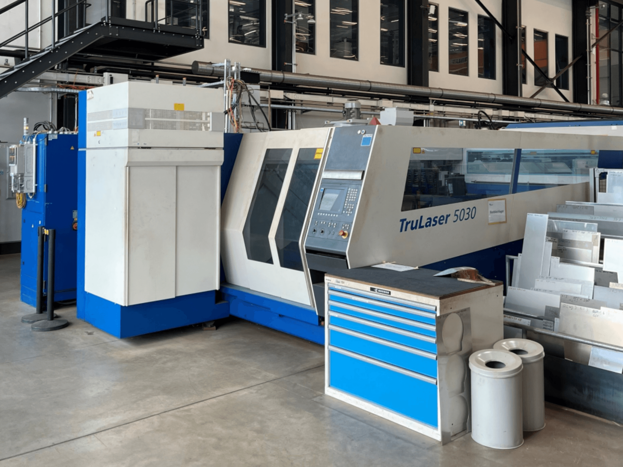 TRUMPF TruLaser 5030 CO2 laser cutting machine in front view, featuring control panel and nearby workbench with metal sheets.