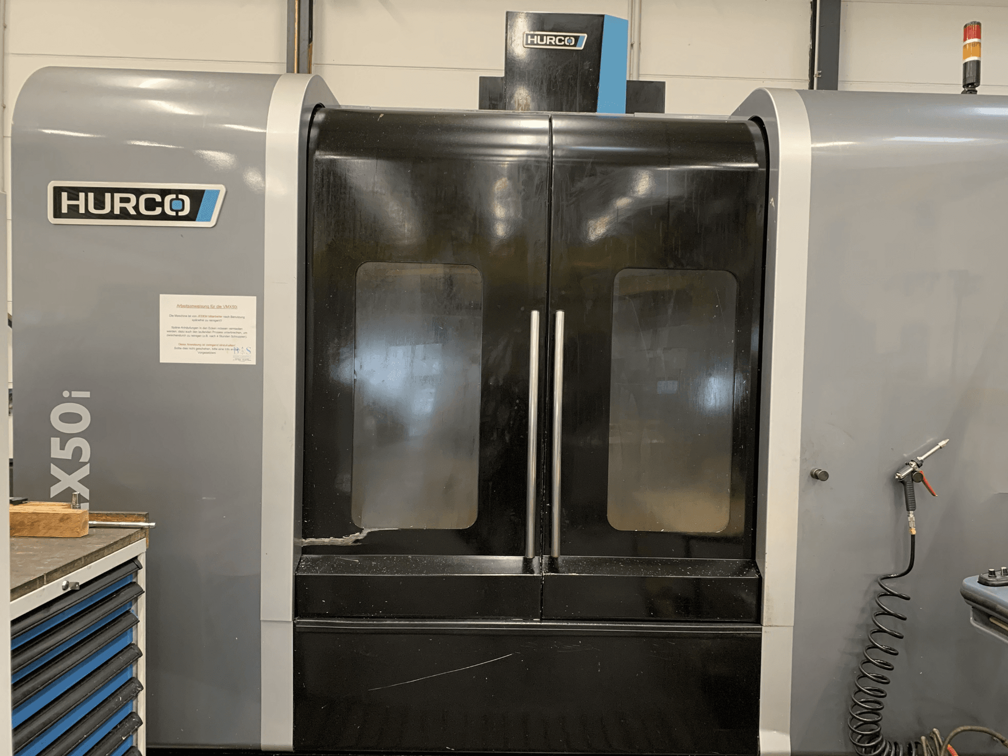 Front view of a Hurco VX50i CNC machine with dual doors, showing technical controls and a maintenance sign on the side.