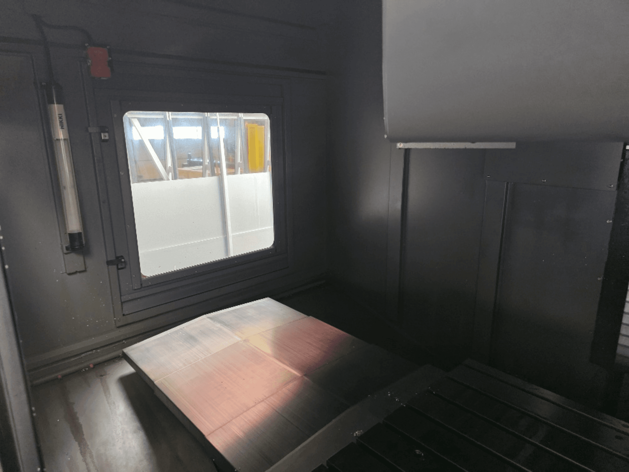 Interior view of a machine enclosure with a polished metal plate, a side window, and minimal lighting. No brand visible.