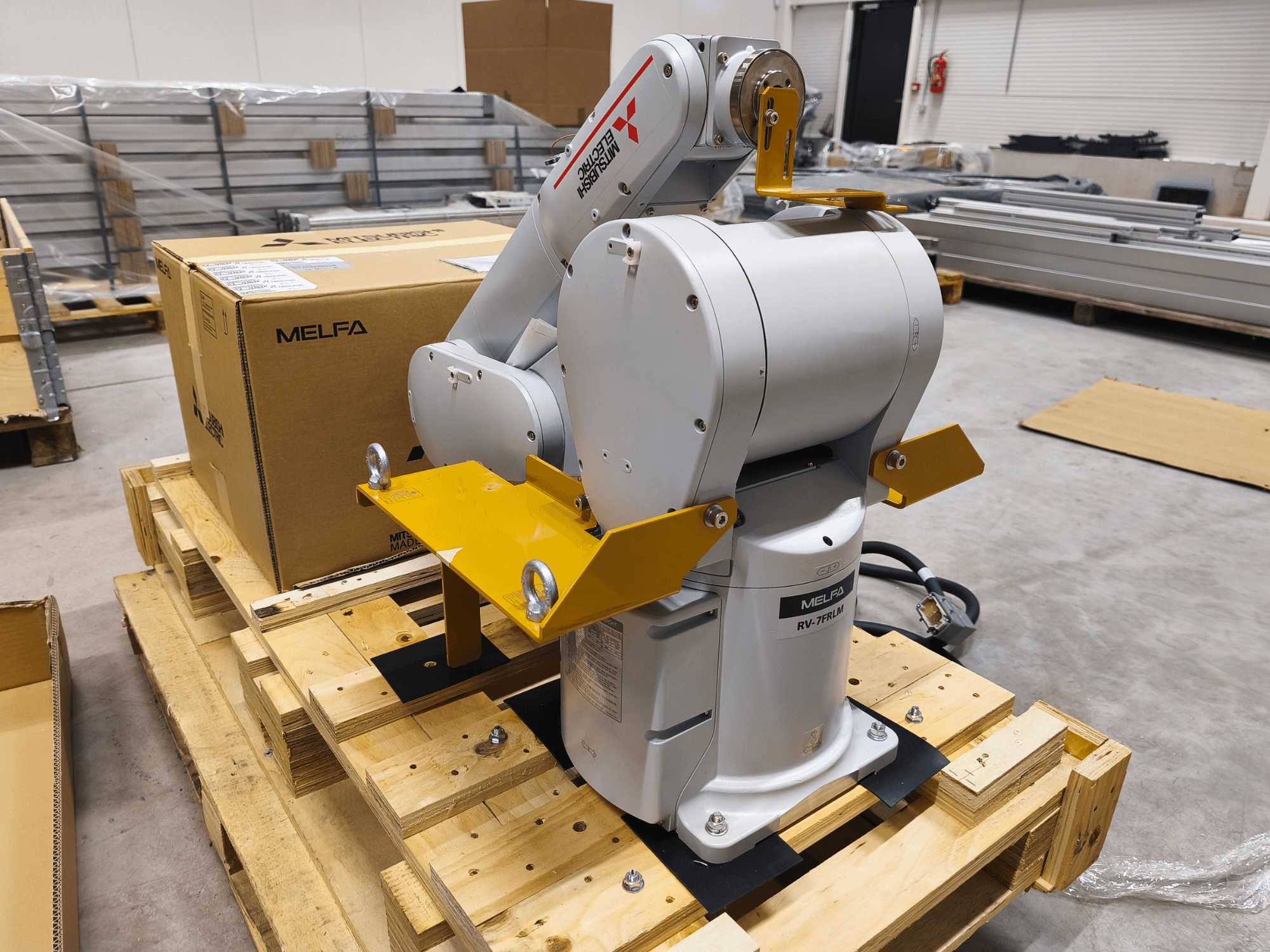 MELFA RV-7FLM industrial robot arm on wooden pallet, front view, with yellow support brackets and gray packing box.