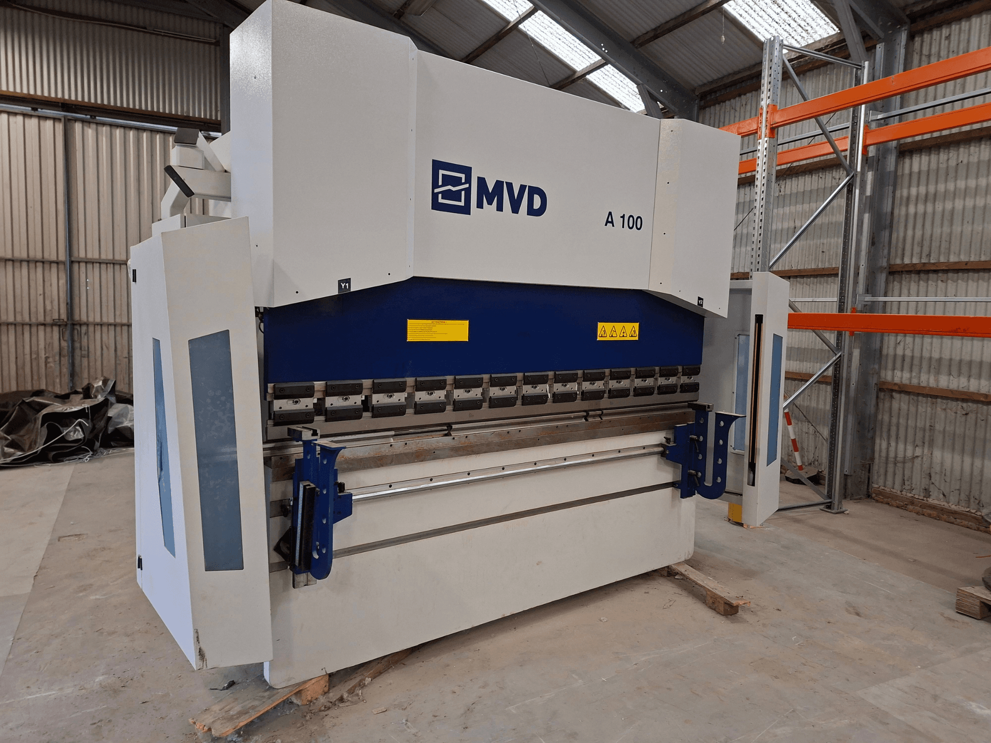 MVD A 100 press brake in a side view, showing controls, safety warnings, and a sturdy metal frame in a spacious workshop.