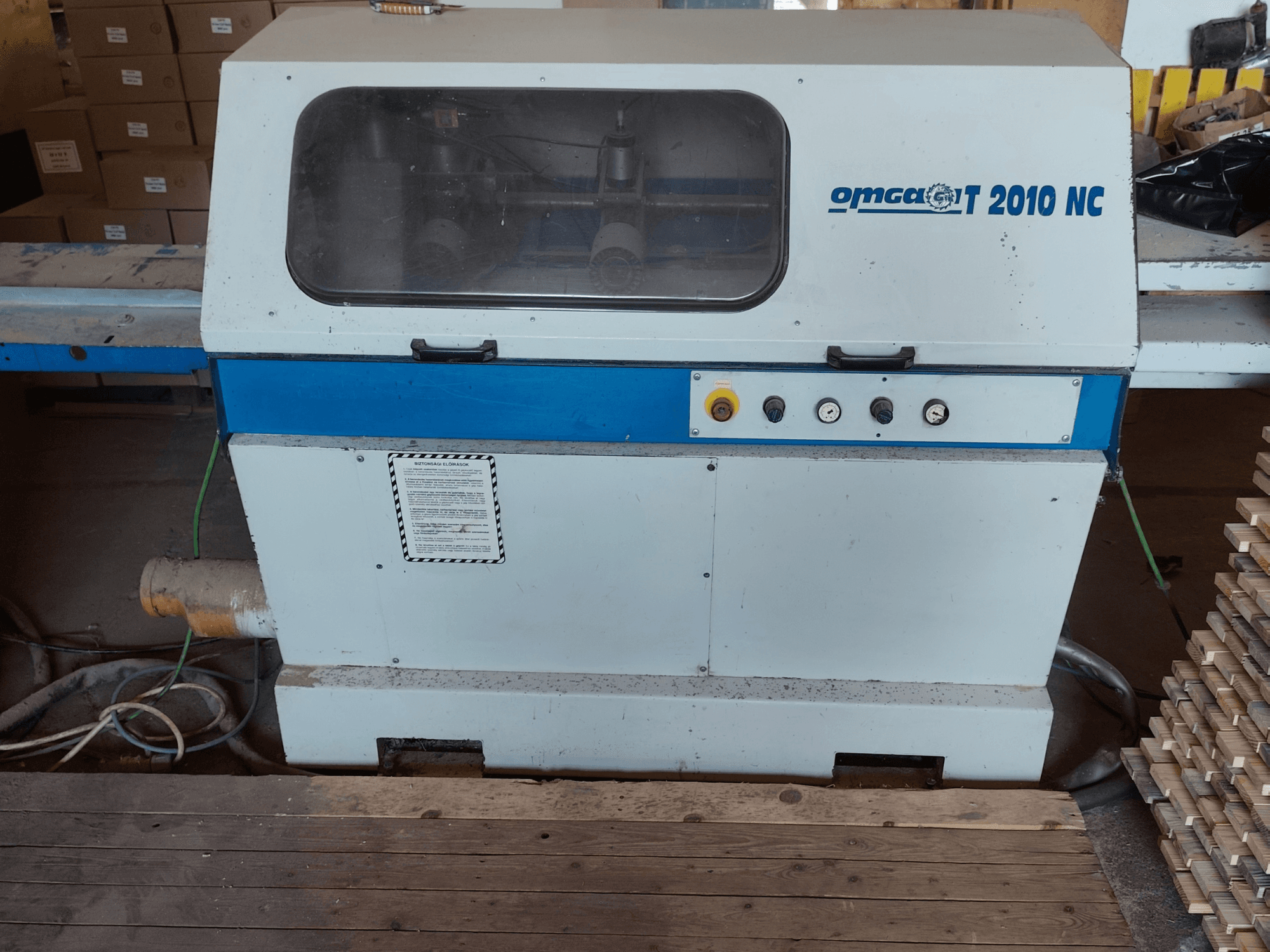 Optimizing Cross-Cut Saw OMGA T 2010 NC, front view, showing control panel and transparent cover, with wood stacked beside it.
