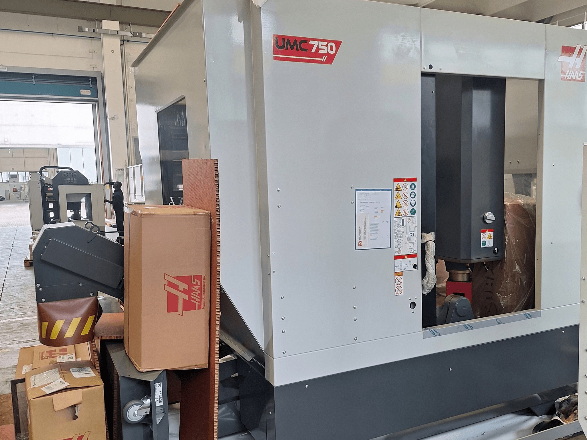 HAAS UMC-750 universal machining center, side view, showcasing control panel and tool storage with warning labels and packaging.