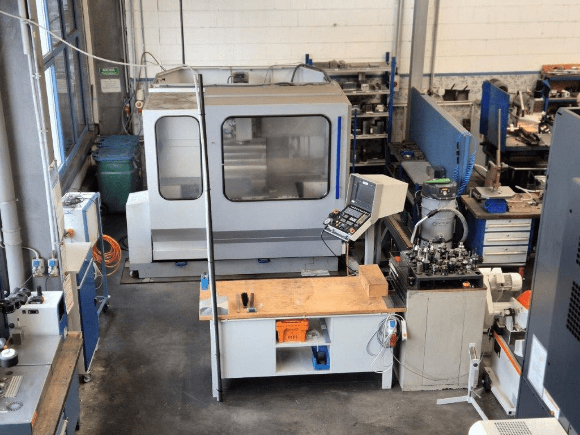 CNC milling machine in a workshop, front view, with control panel, work surface, and tools visible in the background.