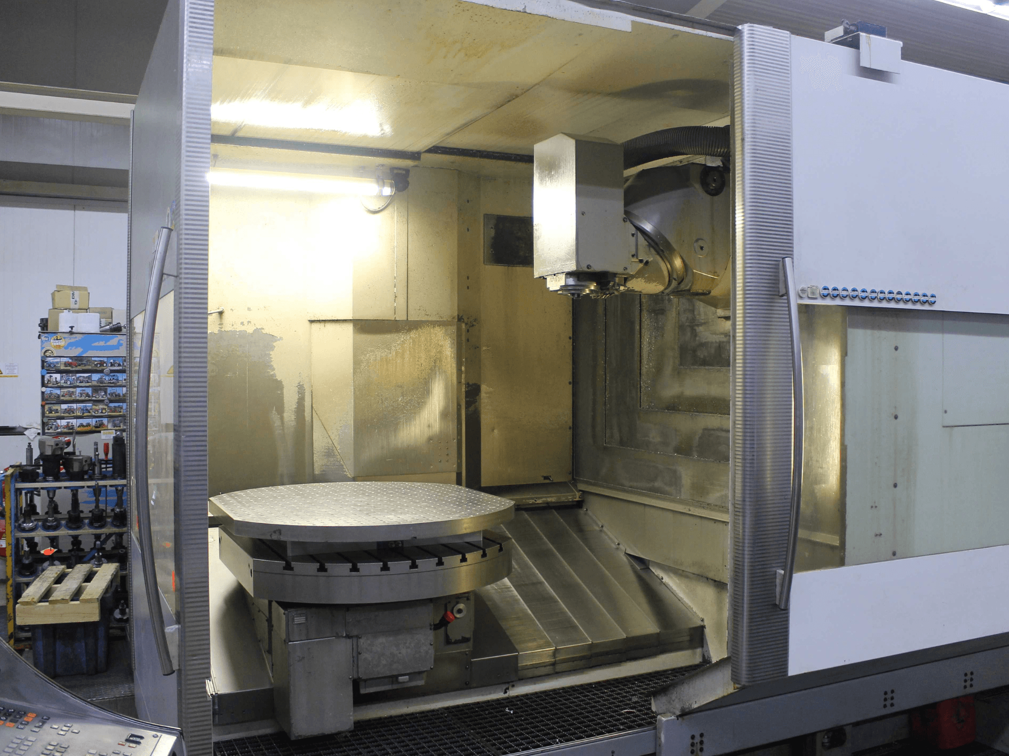 CNC milling machine with a circular rotary table and cutting head, shown from a front view, featuring metallic surfaces and lighting.