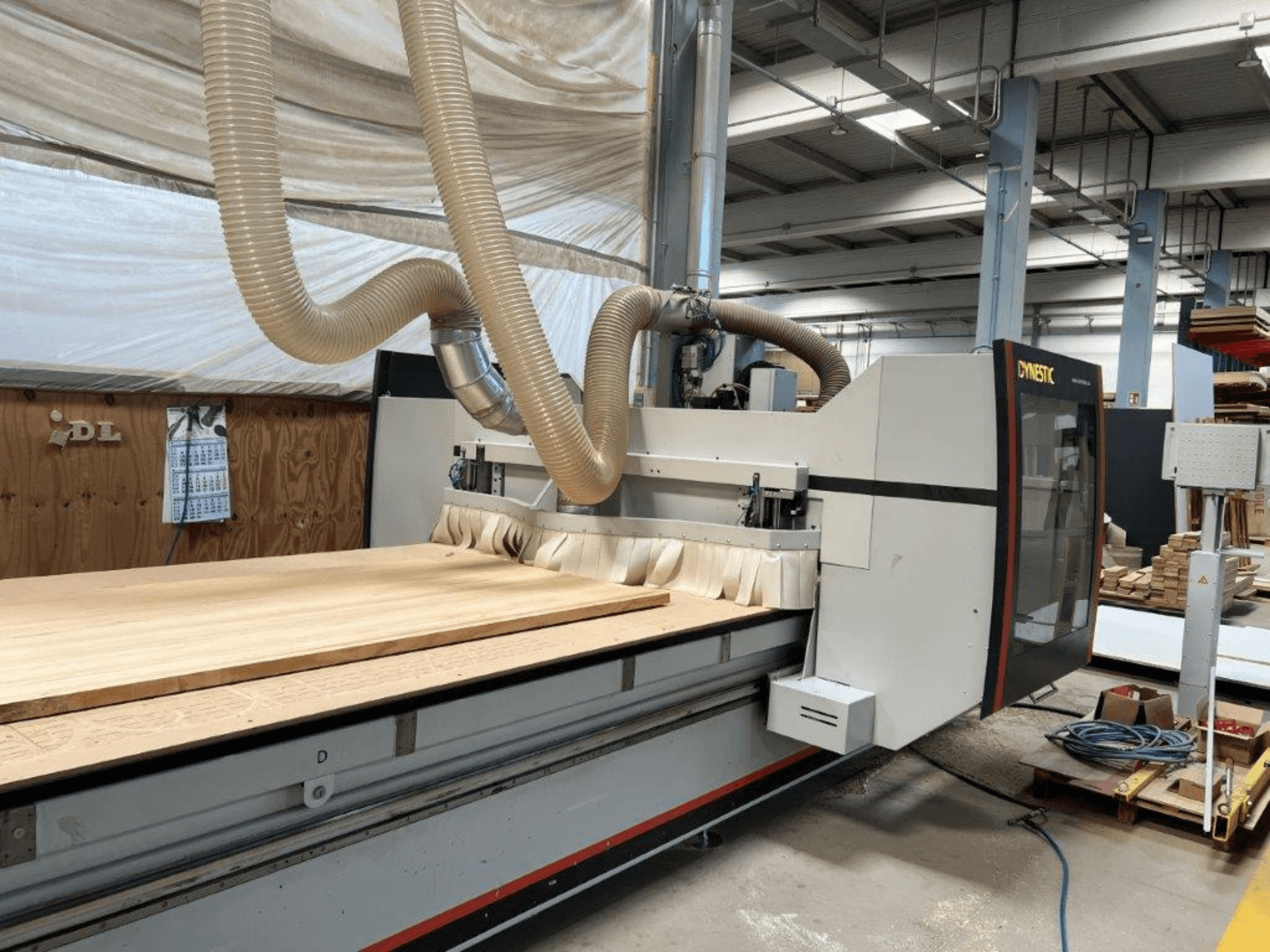 CNC machine by Dynestic, front view, featuring a large wooden workpiece and dust extraction hose in a workshop setting.