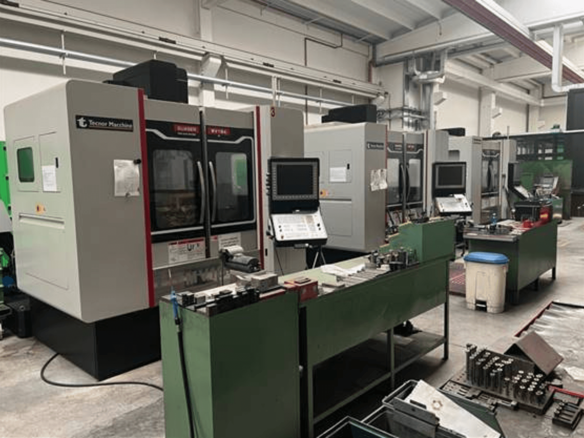 Four CNC machines from Tecnoer Marchine in a workshop, front view, with tools and parts on green workbenches.