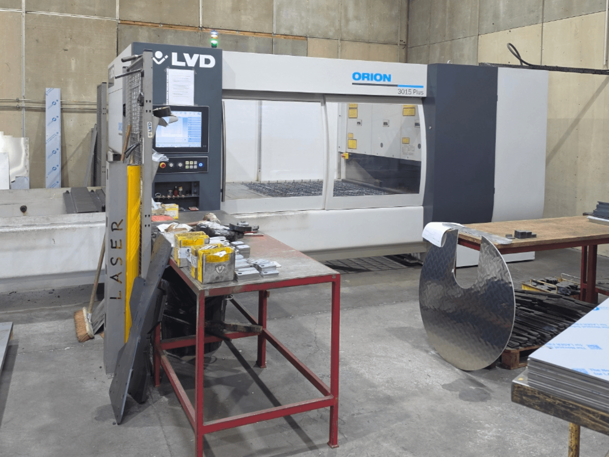 LVD Orion 3015 Plus CO2 laser cutting machine, front view with control panel; surrounded by metal parts on a work table.