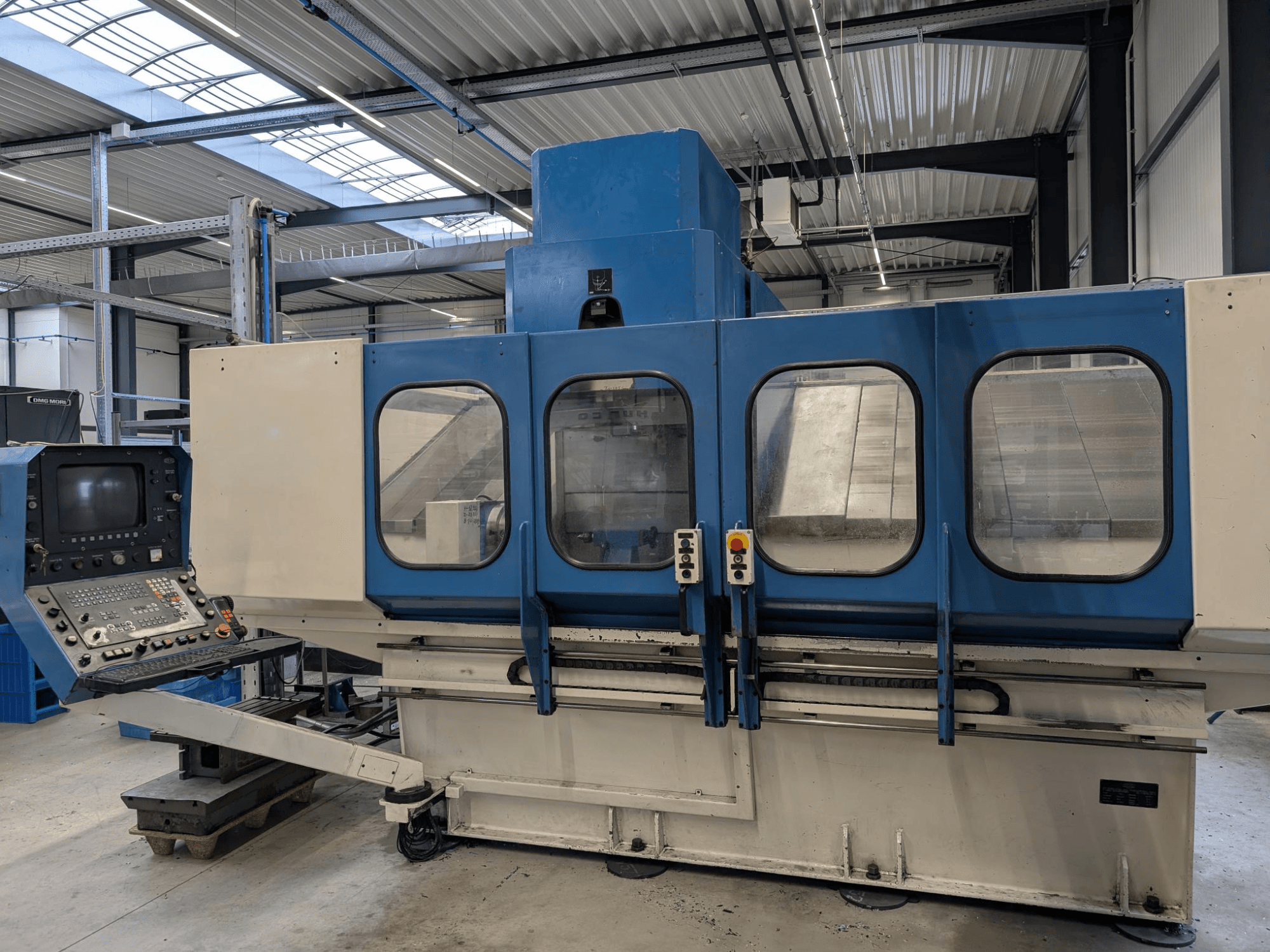Blue industrial CNC machine with control panel, front view. Large glass windows, visible machinery components and buttons.