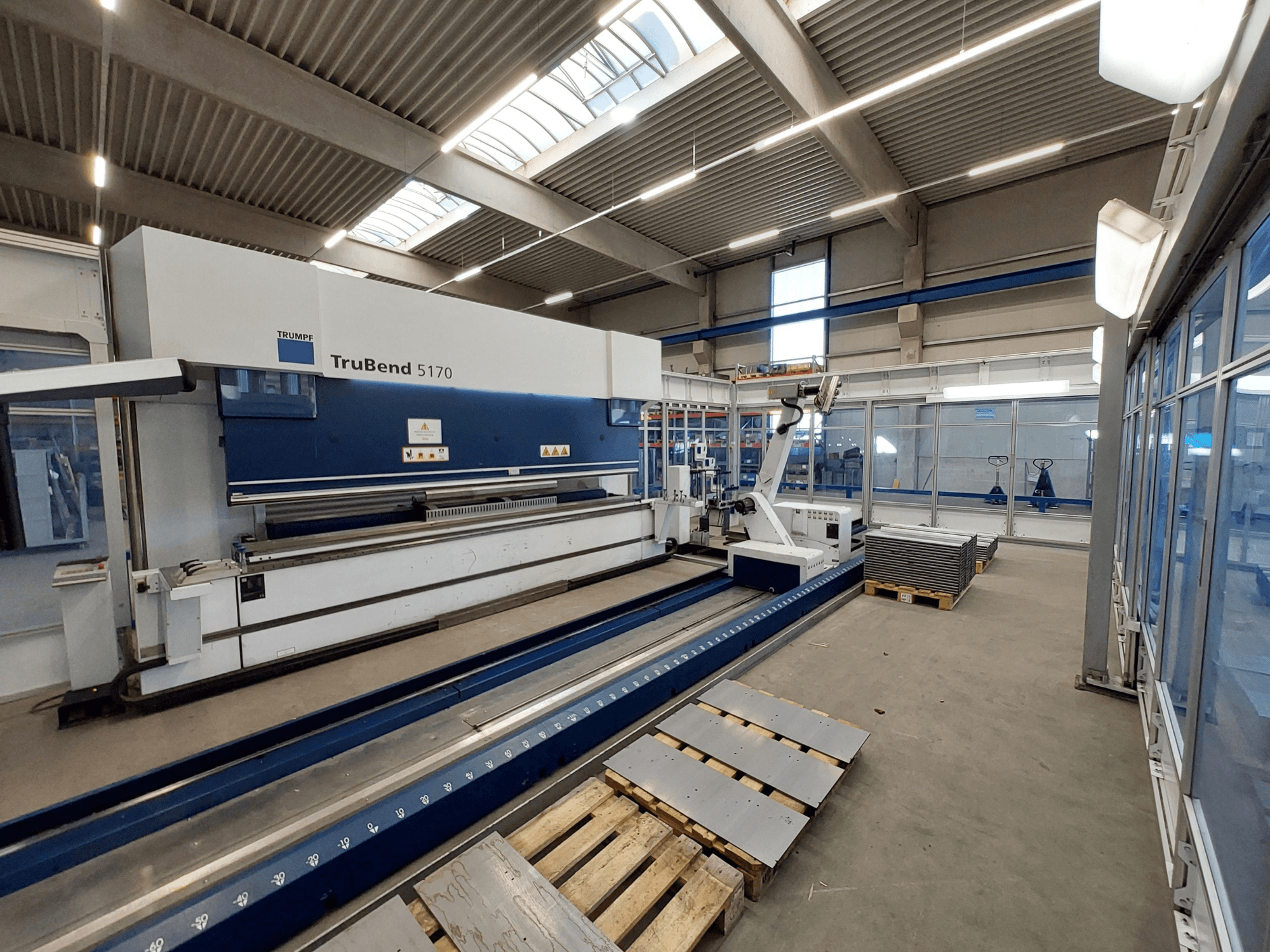 TRUMPF TruBend 5170 press brake in a workshop, side view, showing bending tool and stacked metal sheets on pallets.