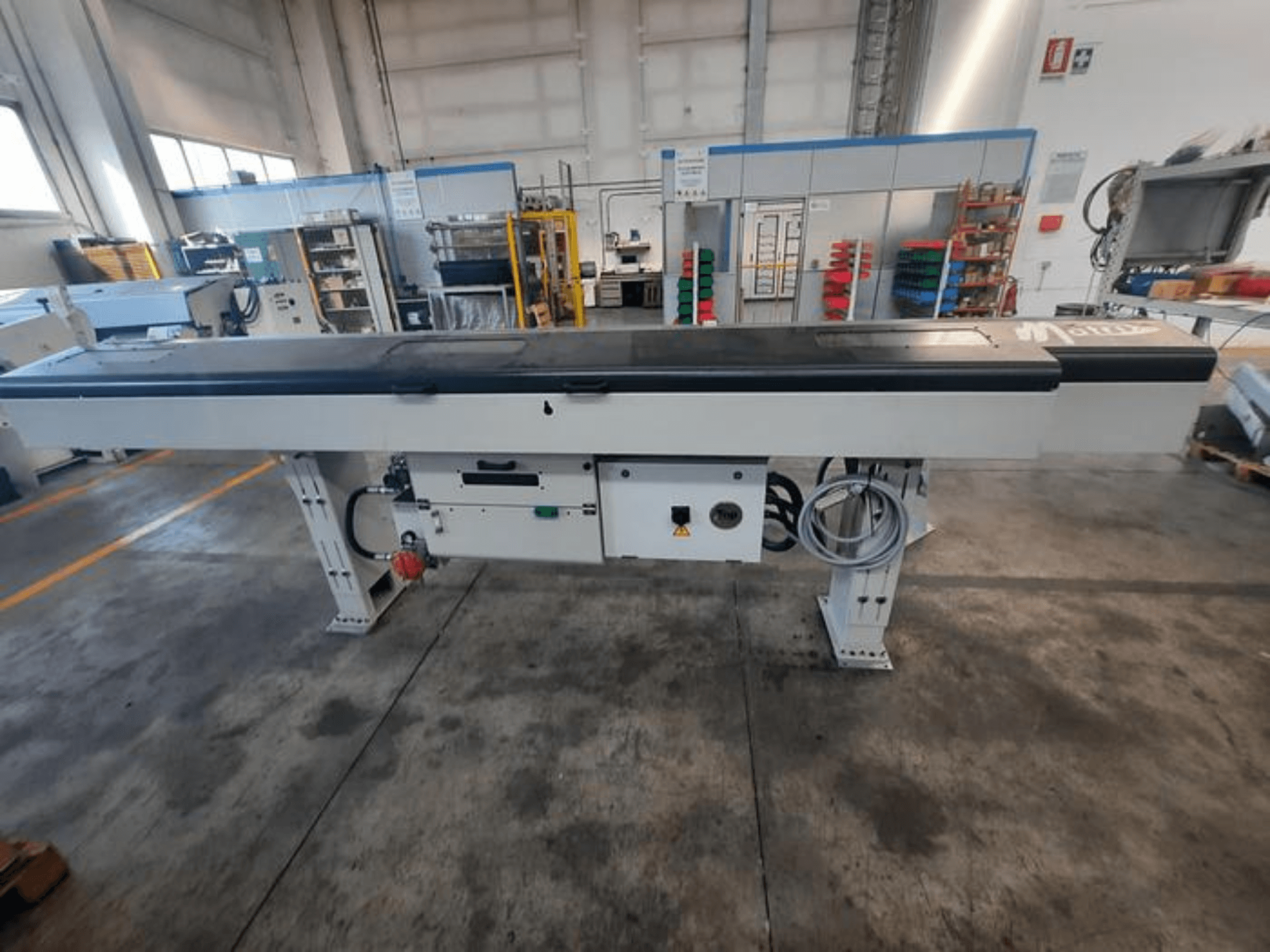 Metal conveyor table with a smooth black top, side view, featuring control buttons and cables; brand logo partially visible.