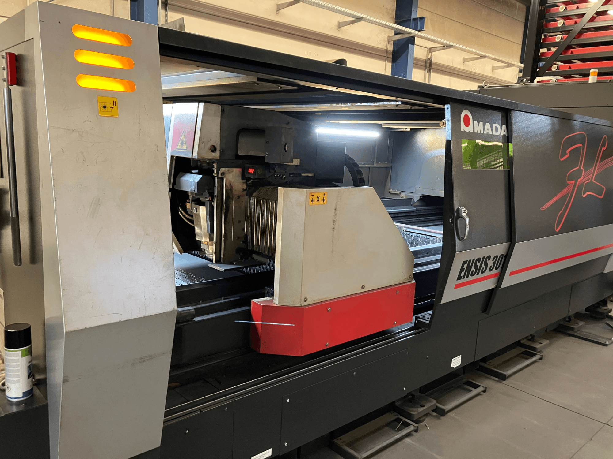 AMADA ENSIS 3015 AJ fiber laser cutting machine, front view, showing cutting head and operating controls inside a metallic frame.