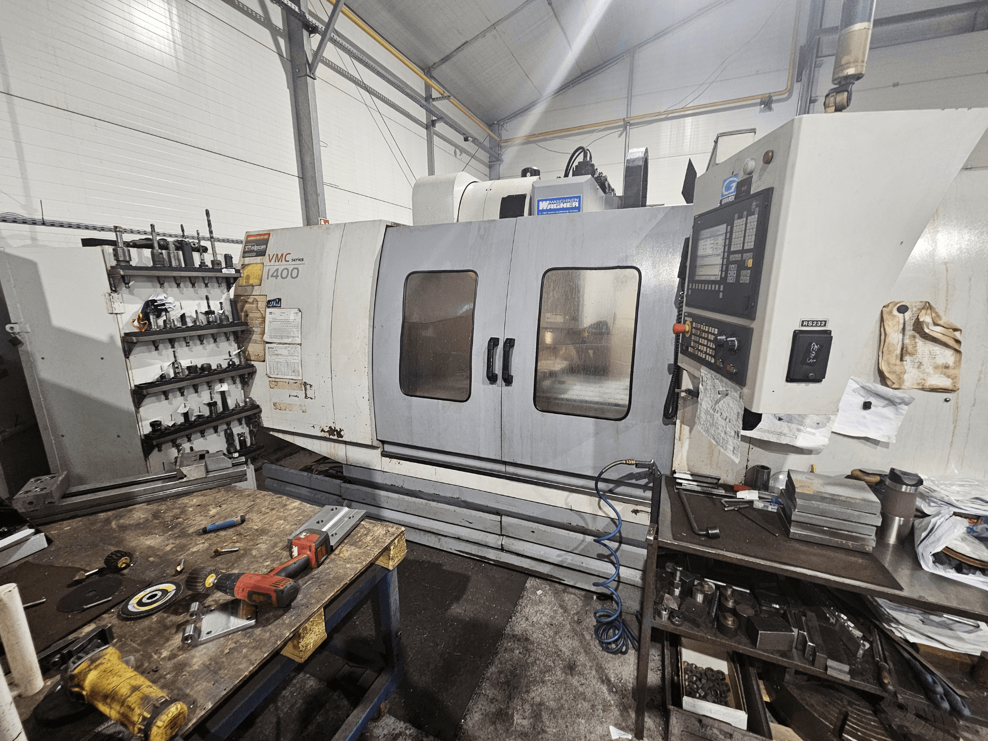 VMC 1400 CNC milling machine in a workshop, front view; tools and materials scattered on the workbench nearby.