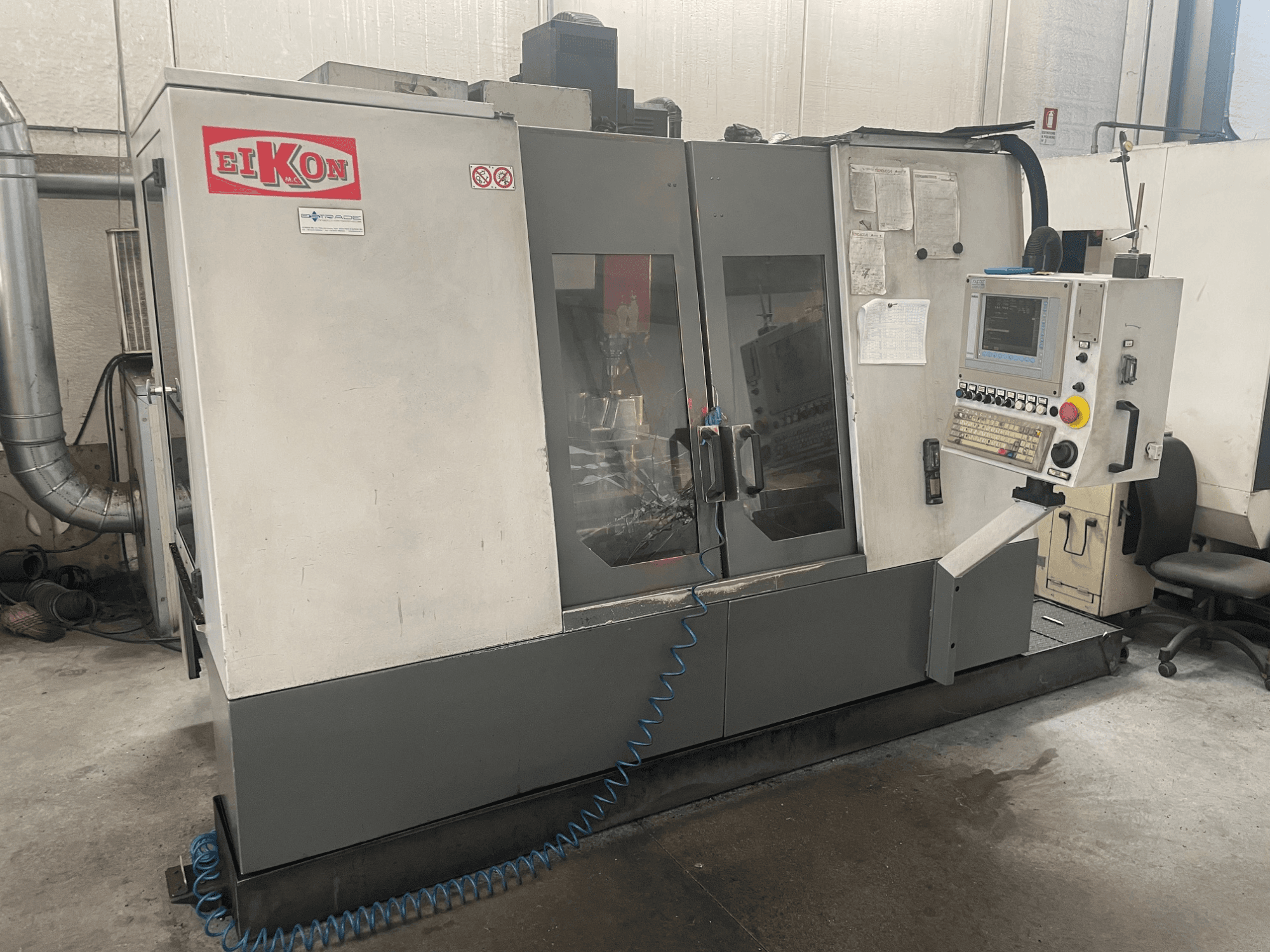 EIKON CNC machine viewed from the front, featuring a control panel, large glass doors, and a blue air hose on the floor.