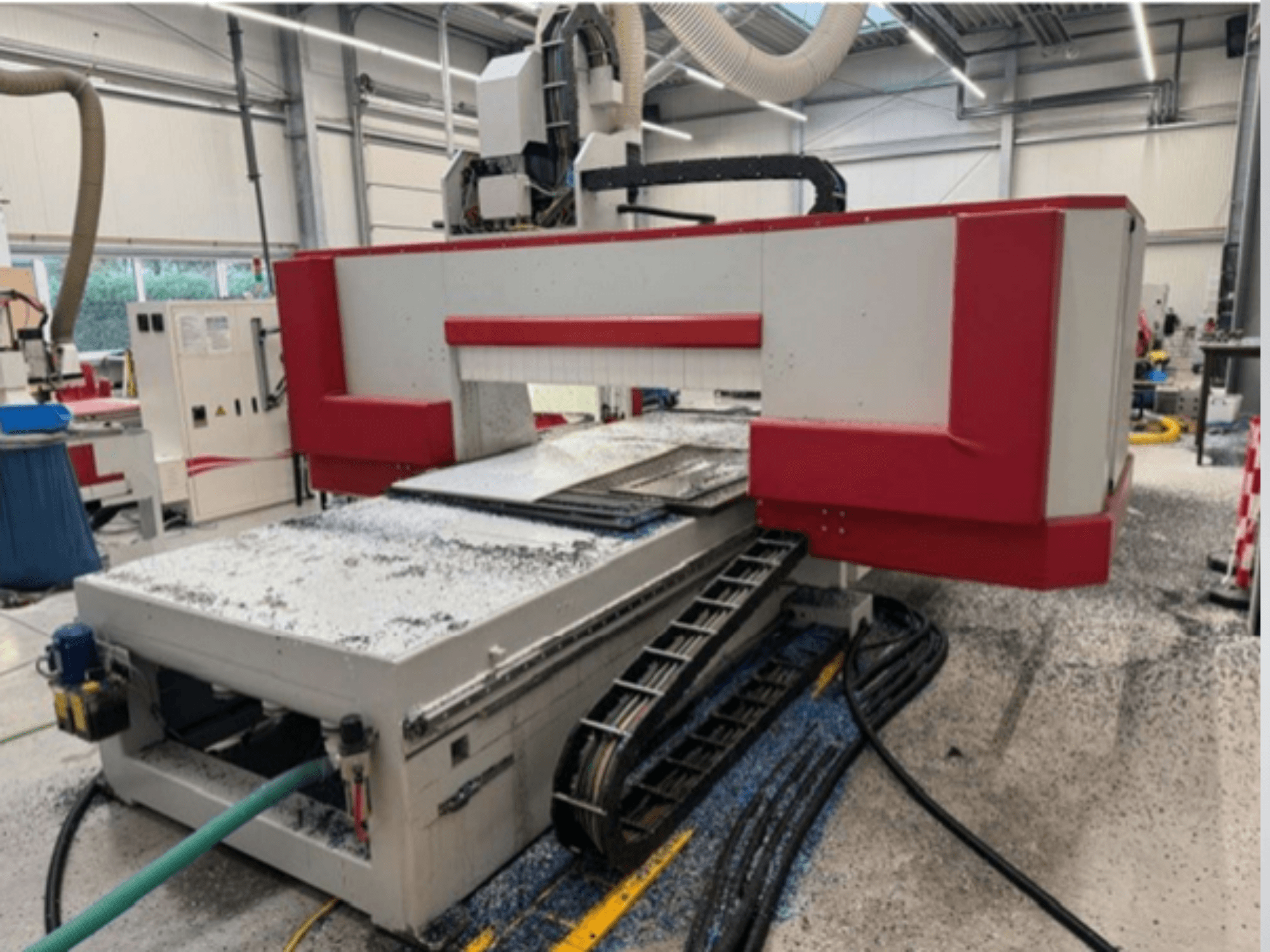 Red and gray CNC machine in a workshop, side view showing material table, coolant hose, and sawdust on the floor.