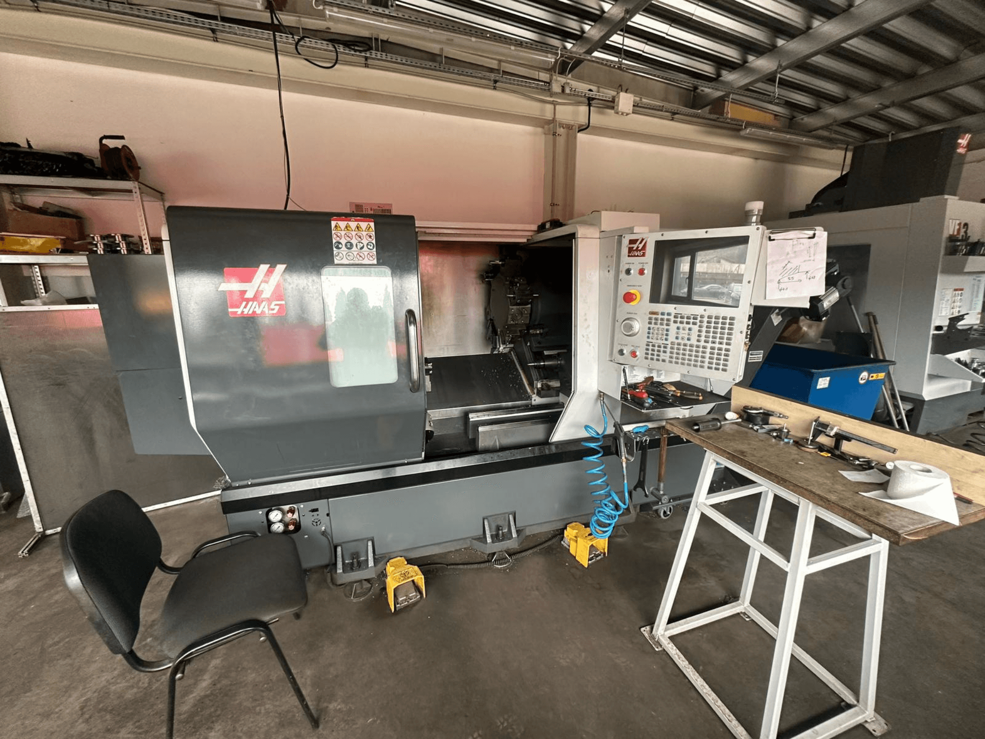 Haas CNC lathe (model ST-10) in a workshop, side view showing controls, tools on a workbench, and a chair nearby.