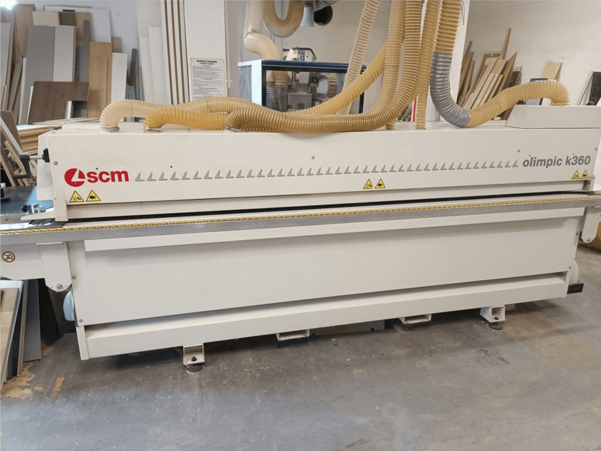 SCM Olimpic K360 T-ER1 edgebander in a workshop, front view showing dust extraction hoses and warning labels.