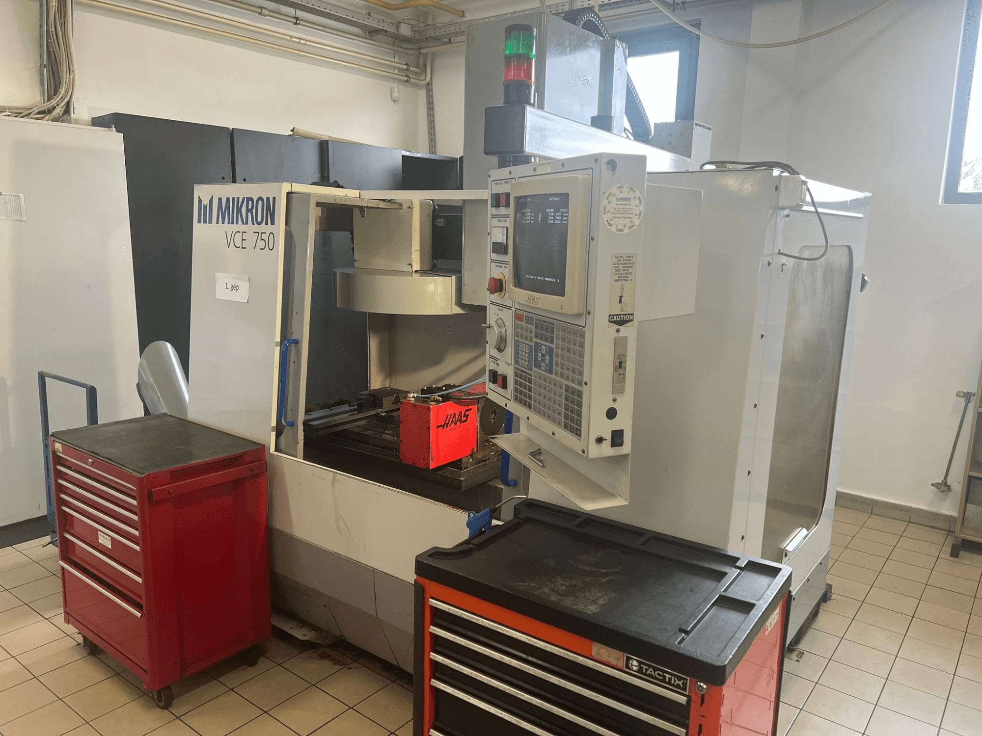 Alt text: "MIKRON VCE 750 Vertical Machining Centre, front view with control panel and red toolboxes, in a spacious workshop."