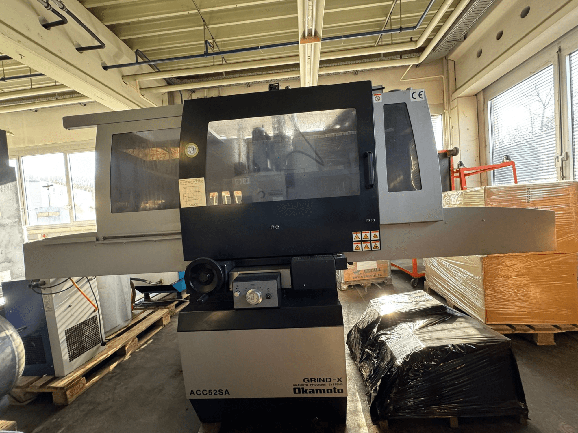 Okamoto ACC 52 SA surface grinding machine in side view, featuring controls and labels, with surrounding pallets and boxes.