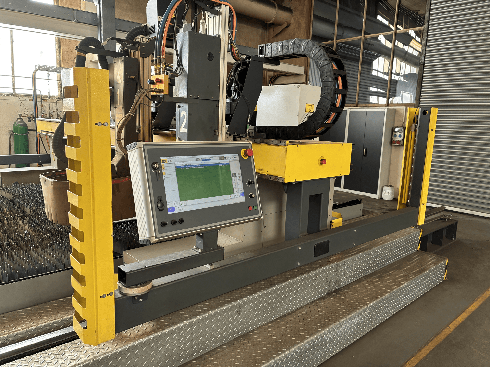 Industrial cutting machine with a touchscreen control panel, yellow safety guard, front view, visible metal grating base.