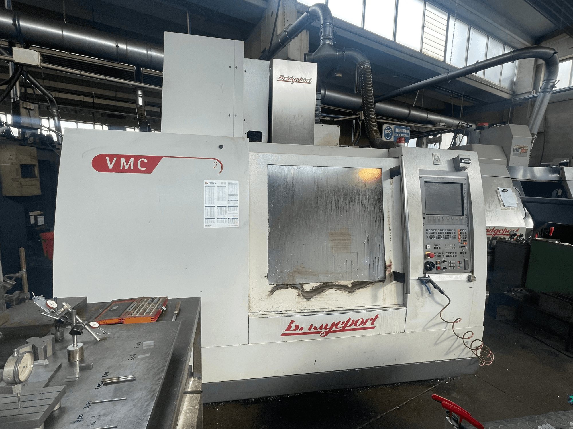 Bridgeport VCM 800-30 vertical machining center in a workshop, featuring a control panel and tool table. Front view with visible details.
