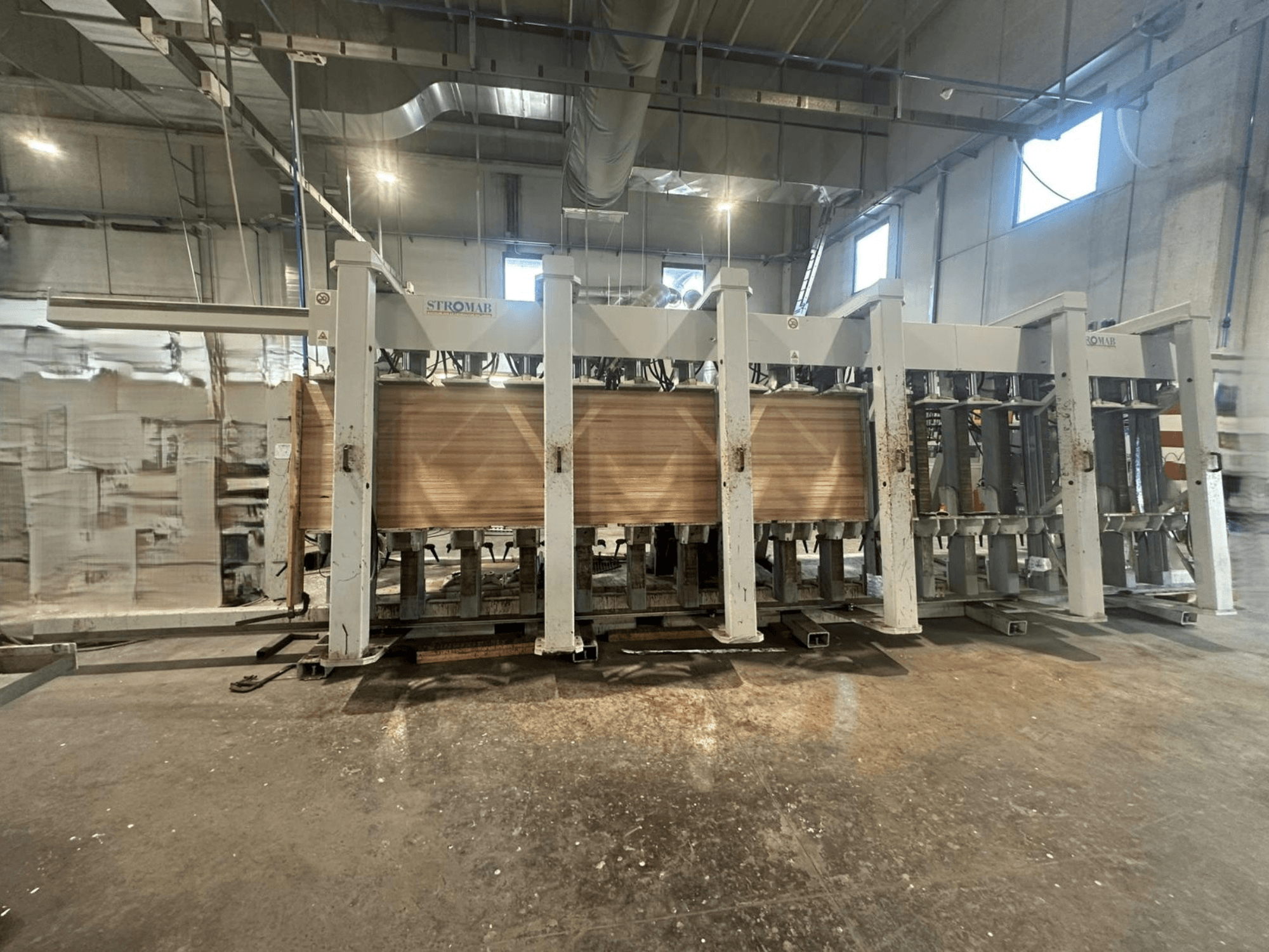 Industrial STROMAB machine in a warehouse, front view, with wooden panels being processed, showcasing metal structure and mechanical parts.