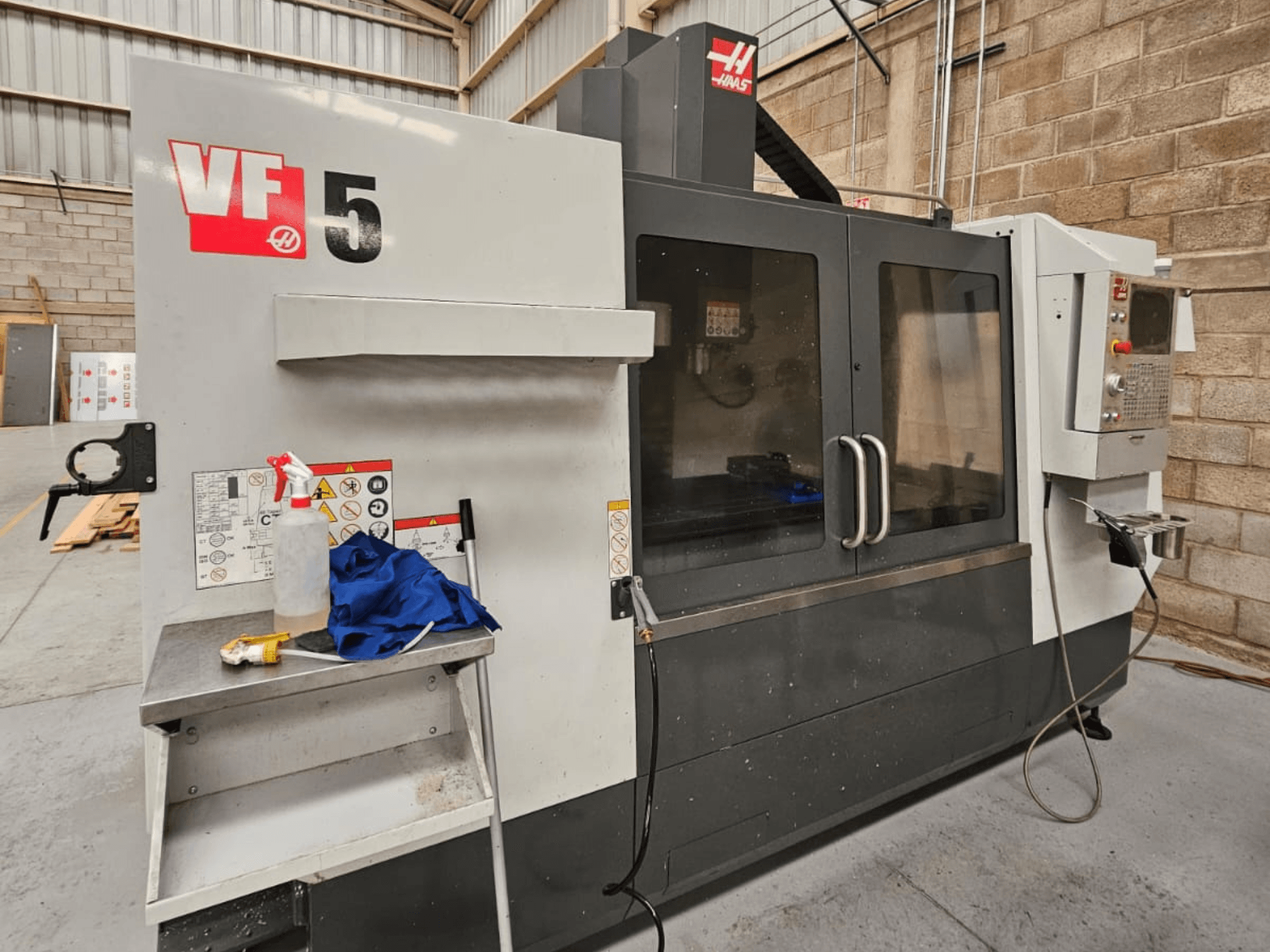 Haas VF-5 CNC milling machine in front view, featuring control panel, coolant bottle, blue cloth, and safety instructions.