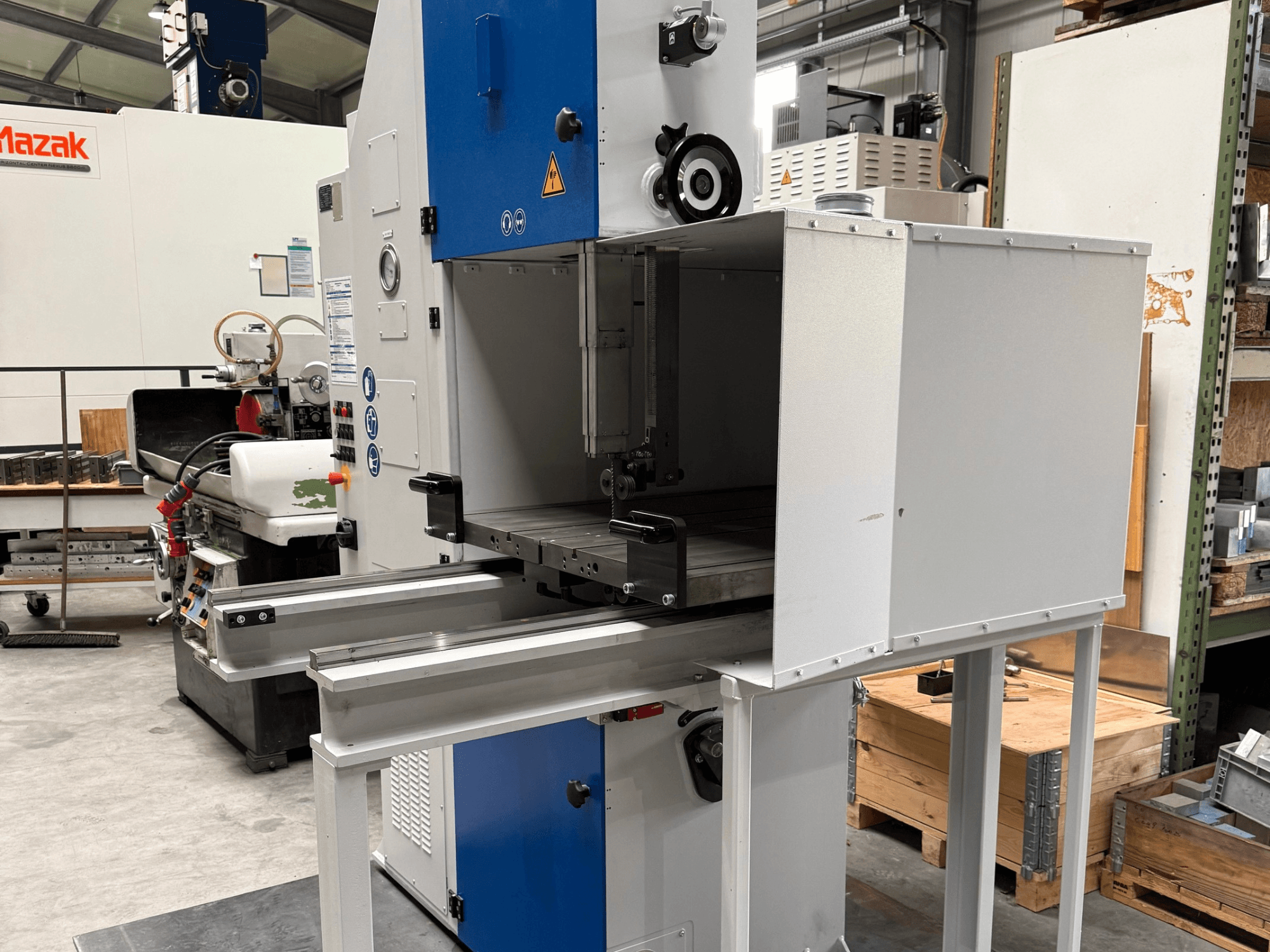 Mazak industrial machine viewed from the front, featuring a blue control panel, metal table, and safety warnings.