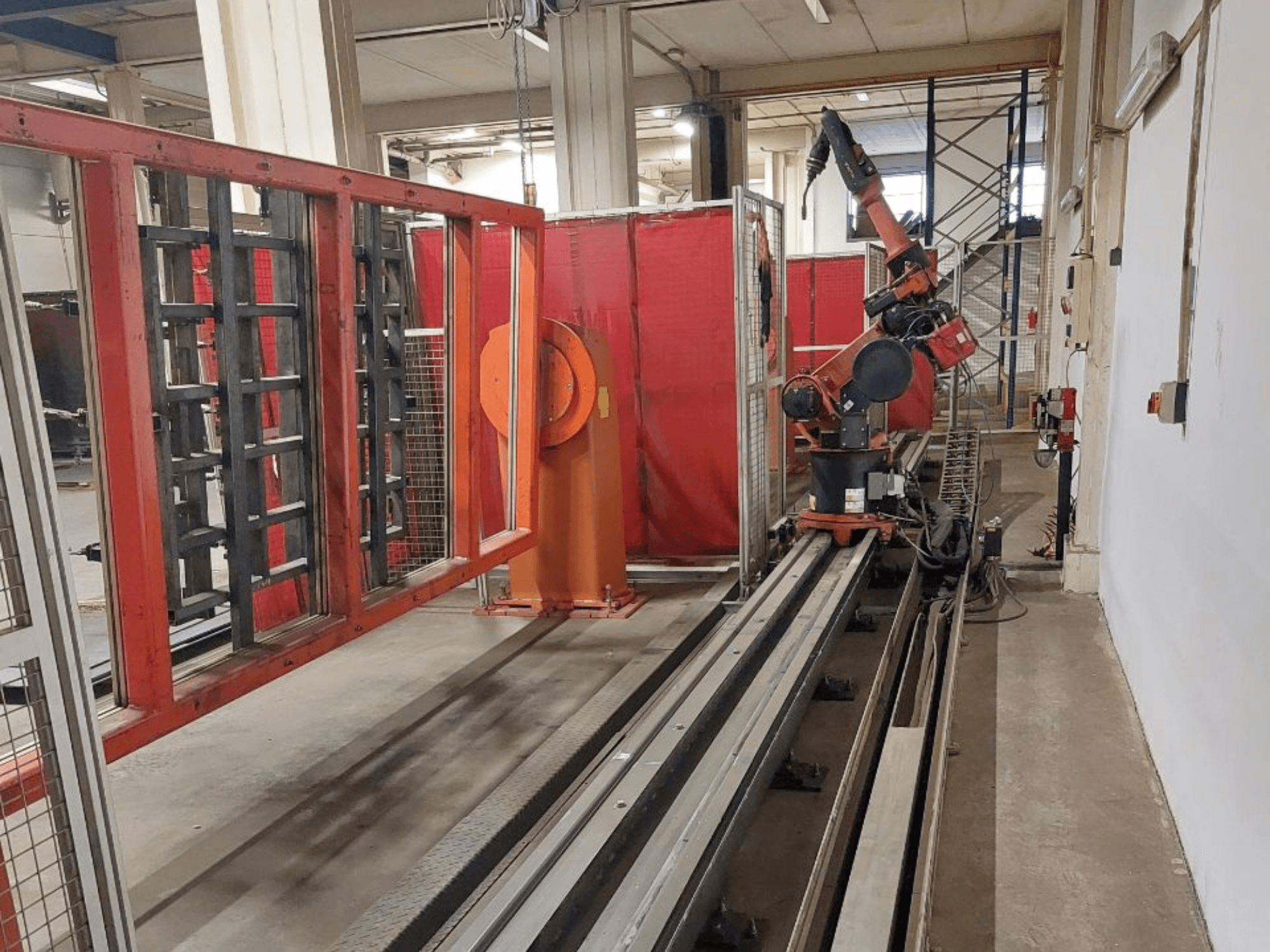 Industrial robotic arm with orange and black colors positioned on metal tracks, surrounded by red protective barriers, front view.