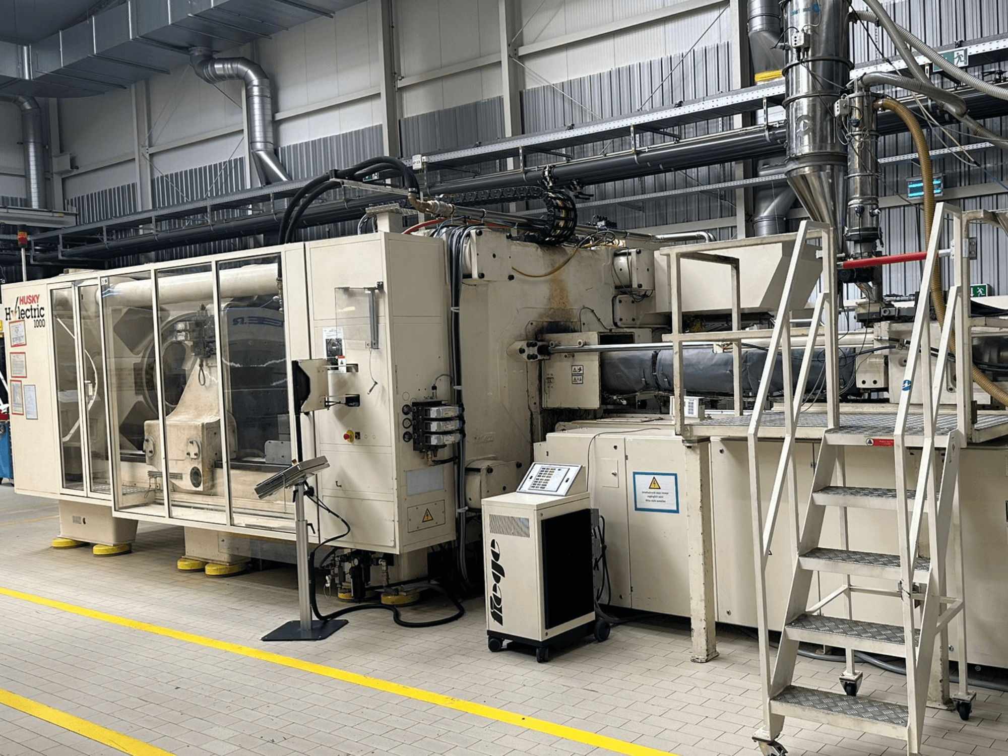 Front view of Husky Hylelectric H1000 RS95/80 electrical injection moulding machine in a factory setting, featuring control panel and steps.