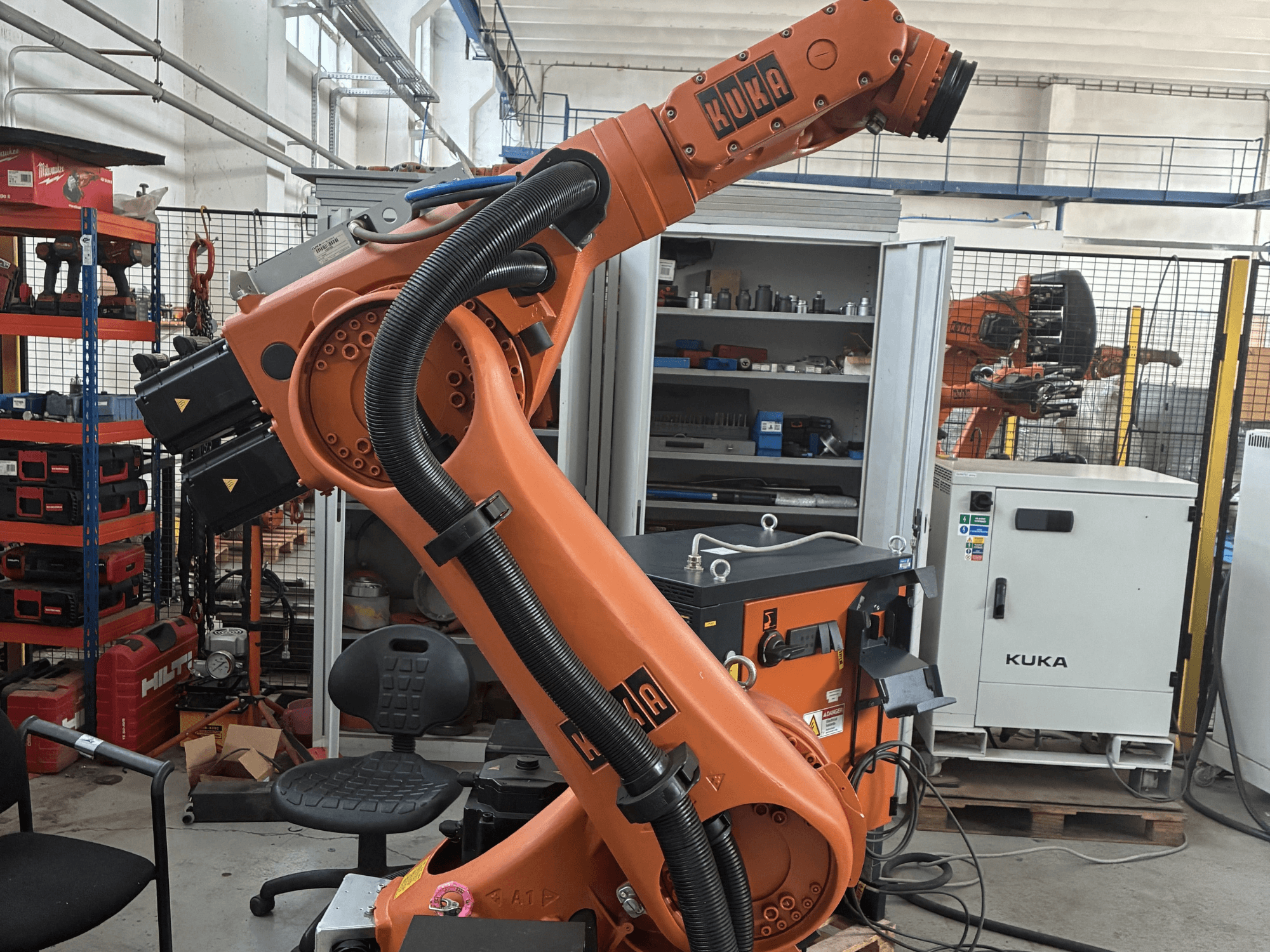 KUKA KR30-3 robot arm in an industrial setting, side view showing cables and controls, with tools and shelving in background.