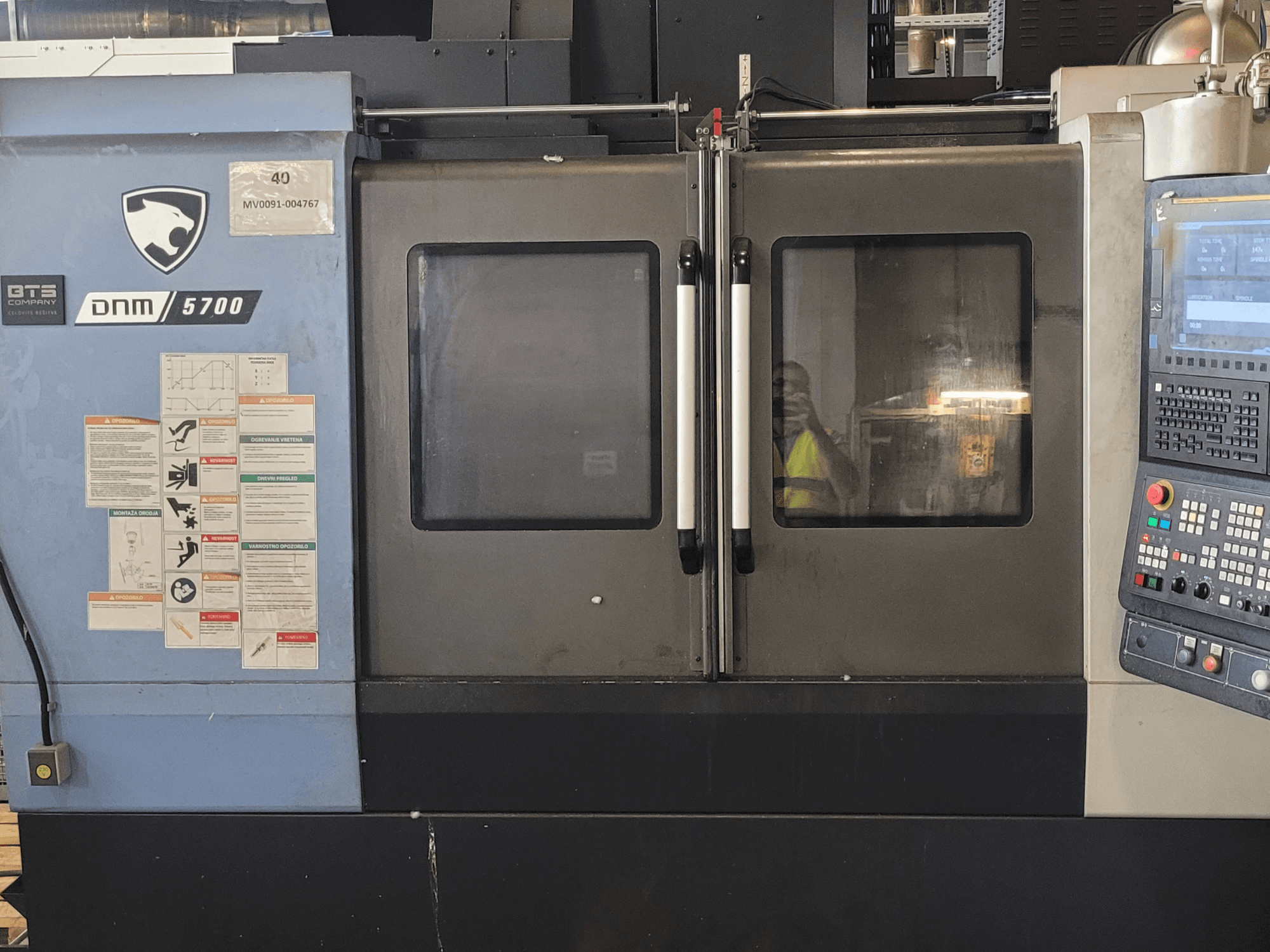 DOOSAN DNM 5700 vertical machining center front view with control panel, safety labels, and doors slightly ajar.