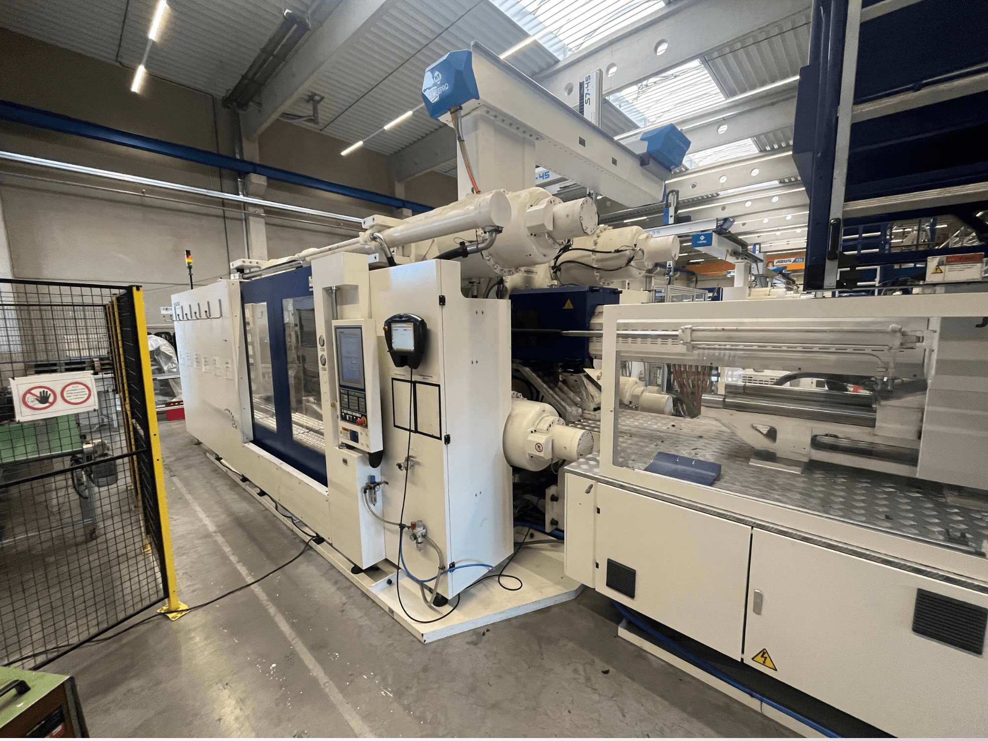 Injection molding machine in white and blue, front view, featuring control panel, robotic arm, and safety signage.