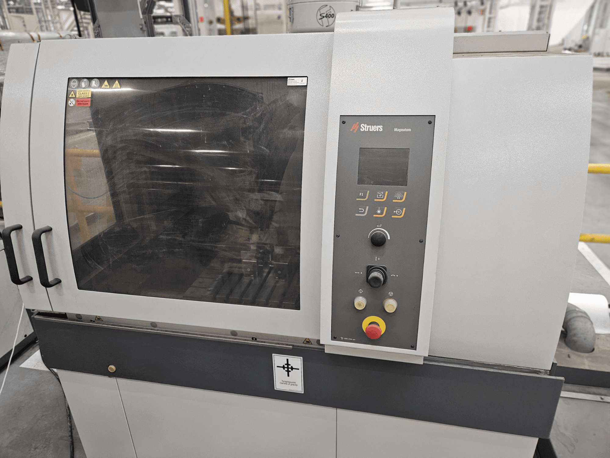 Struers Magpentor machine front view with control panel, buttons, and a large viewing window. Technical labels visible.