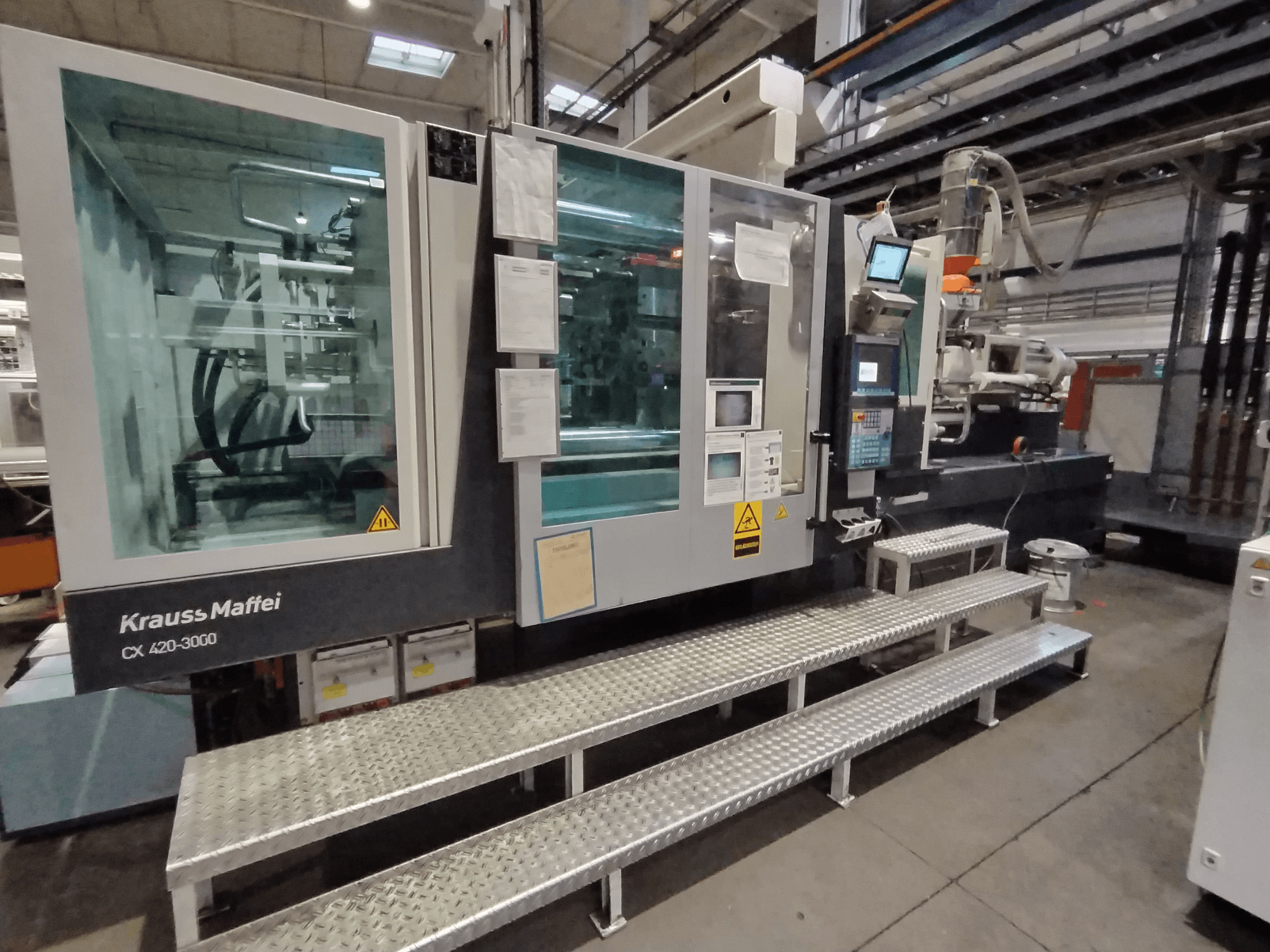 Krauss Maffei CX 420-3000 injection molding machine, side view, with control panel and safety signage visible, set on metal steps.