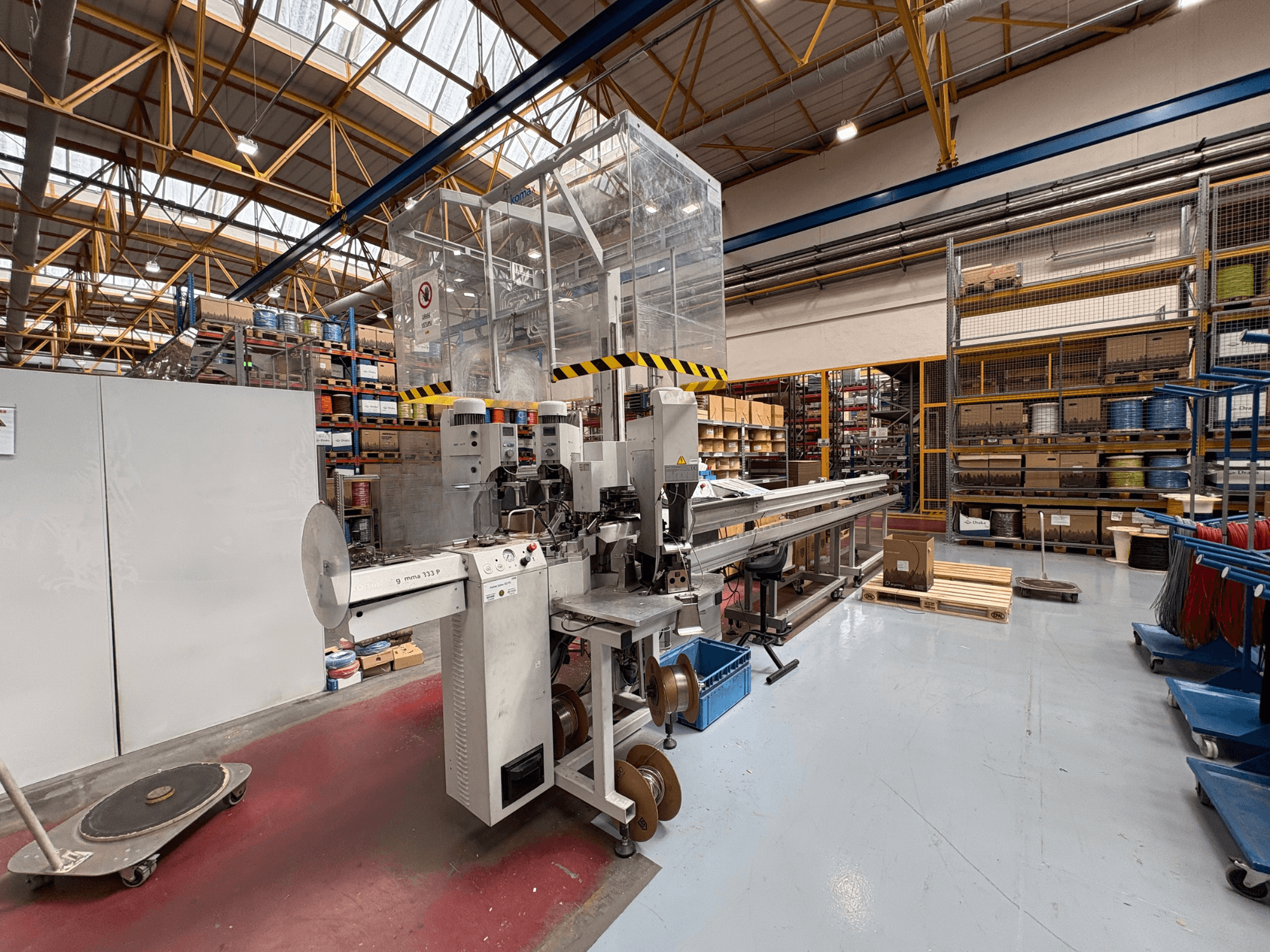 KOMAX GAMMA 333 PC machine tool in a warehouse, side view showing controls, conveyor system, and surrounding shelves.