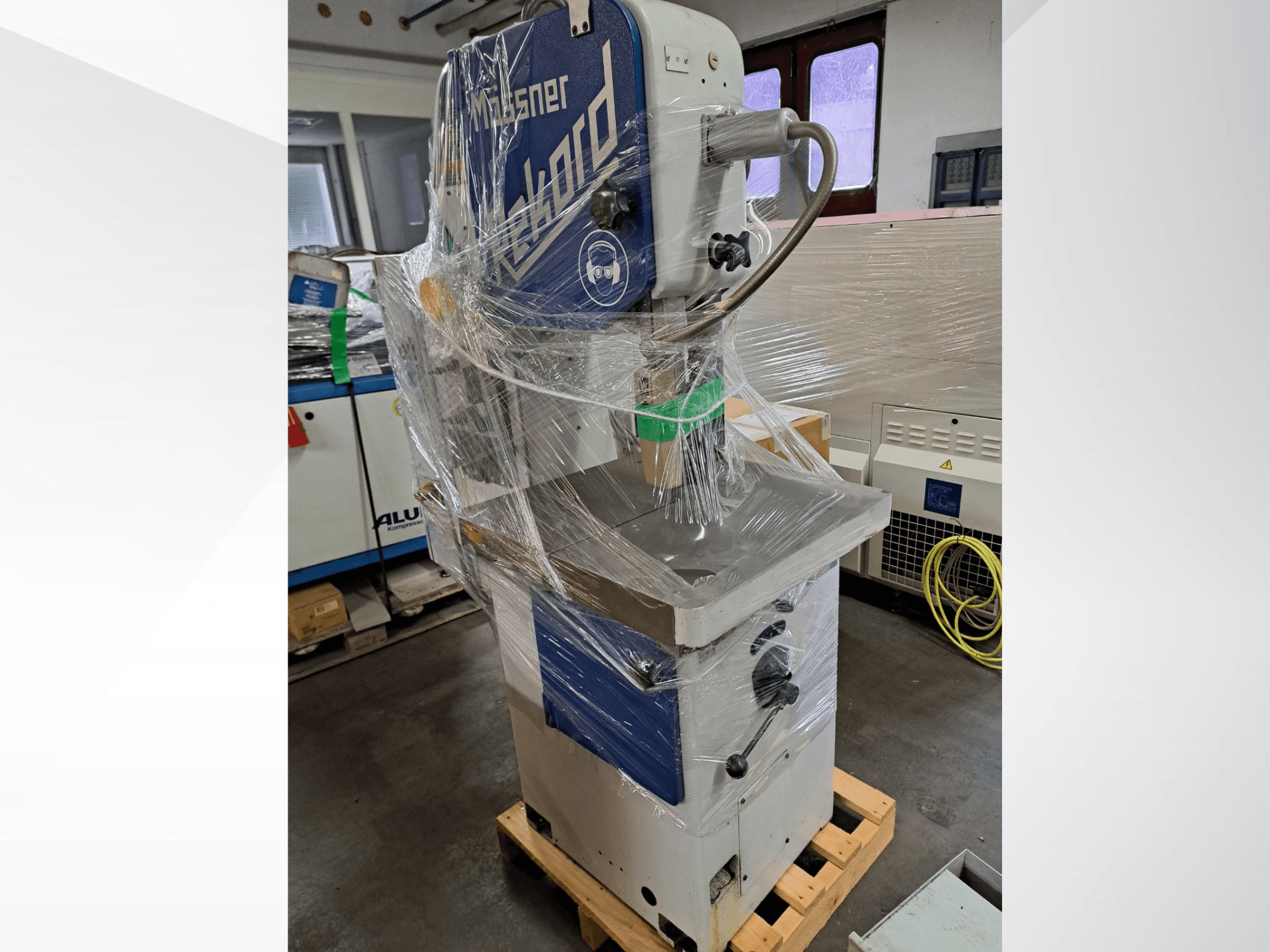 MOSSNER Rekord metal band saw wrapped in plastic, side view showing control panel and base on a wooden pallet in a workshop.